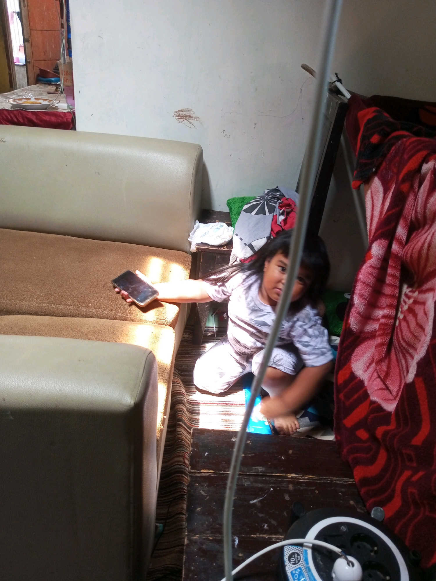 A young girl with long dark hair is crouching on a wooden floor, holding a dark smartphone in her right hand. She is wearing a light-colored shirt with a gray pattern and matching pants. Sunlight streams in from the left, casting shadows. Behind her is a couch with beige and brown upholstery. To the right, a red blanket with a floral pattern is draped over furniture. In the foreground, a power strip with multiple outlets and a white cord is visible on the floor. The scene appears to be indoors in a room with light-colored walls. The location is identified as Krasak, Indonesia.