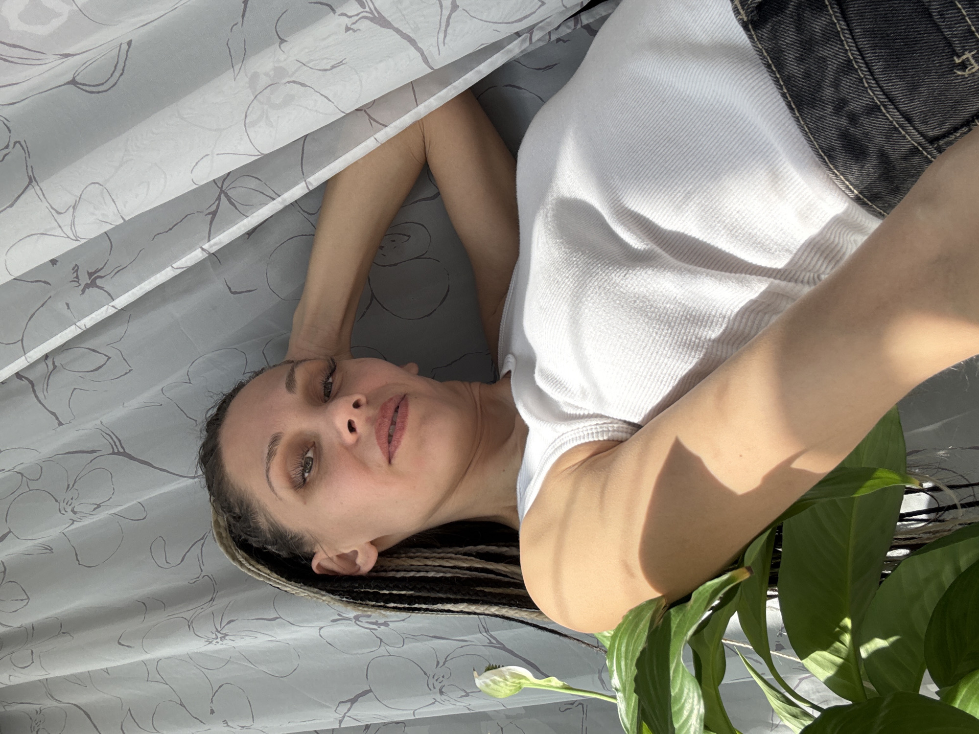 A woman with braided hair lies on a patterned surface, possibly a bed, next to a sheer curtain with a floral pattern. She is wearing a white tank top and what appears to be a dark-colored denim jacket or shorts. A peace lily plant is visible in the foreground. The image is taken at an angle, creating a slightly disorienting perspective. There is no information in the image to determine that the image was taken in Rusanovo, Russia.