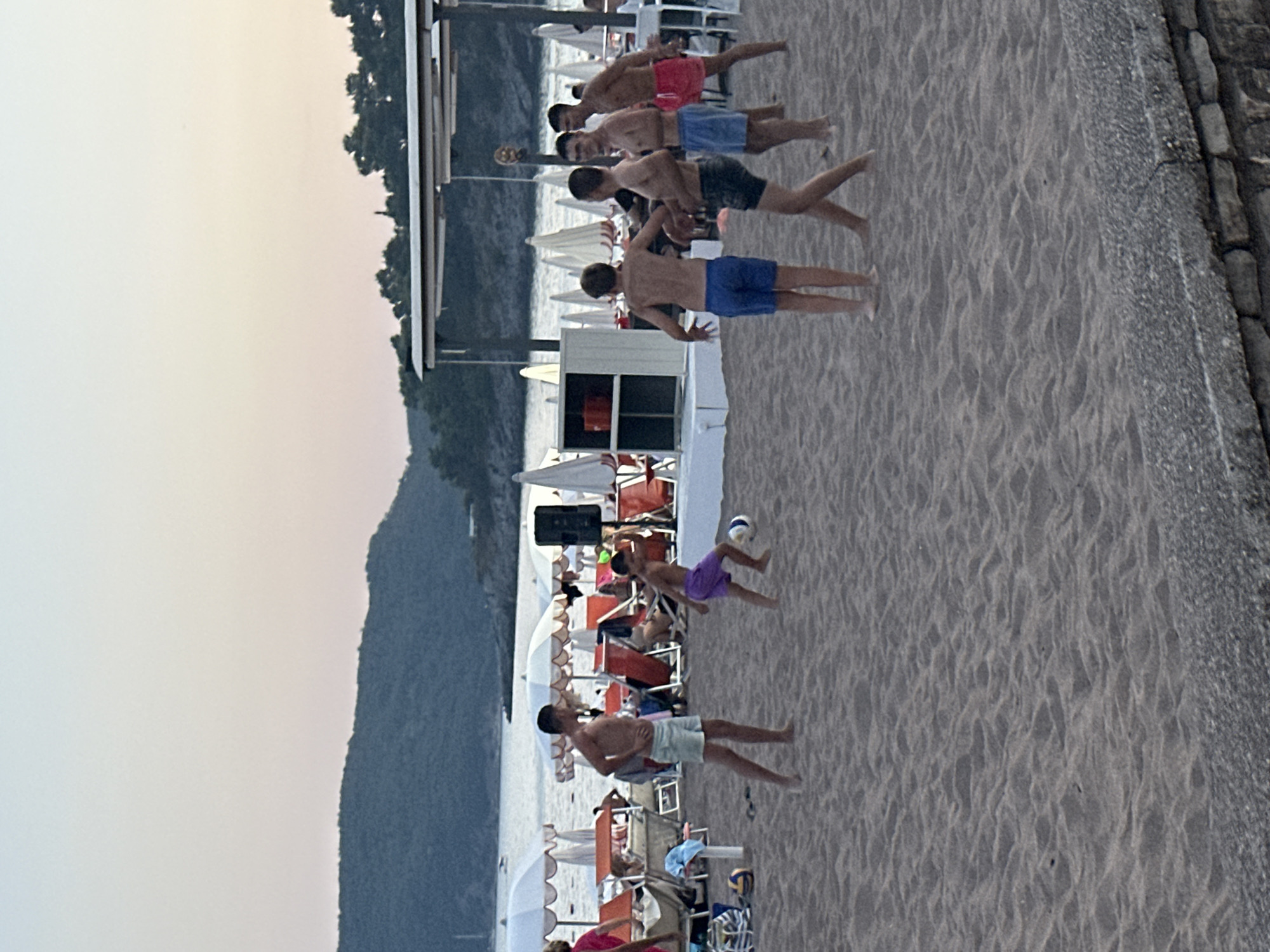 In the city of Boreti, Montenegro, on a sandy beach, a group of people are playing soccer. The scene unfolds during twilight, with a pale sky and a gentle sea in the background. Several individuals, including children and adults, are clad in swimwear, actively engaged in a game of soccer. Some are barefoot, while others wear sandals. Beach umbrellas and lounge chairs are visible in the background, hinting at a recreational setting. The sand is marked with footprints and slight undulations, indicating recent activity. A stone wall borders the lower edge of the frame, separating the beach from an unseen area.