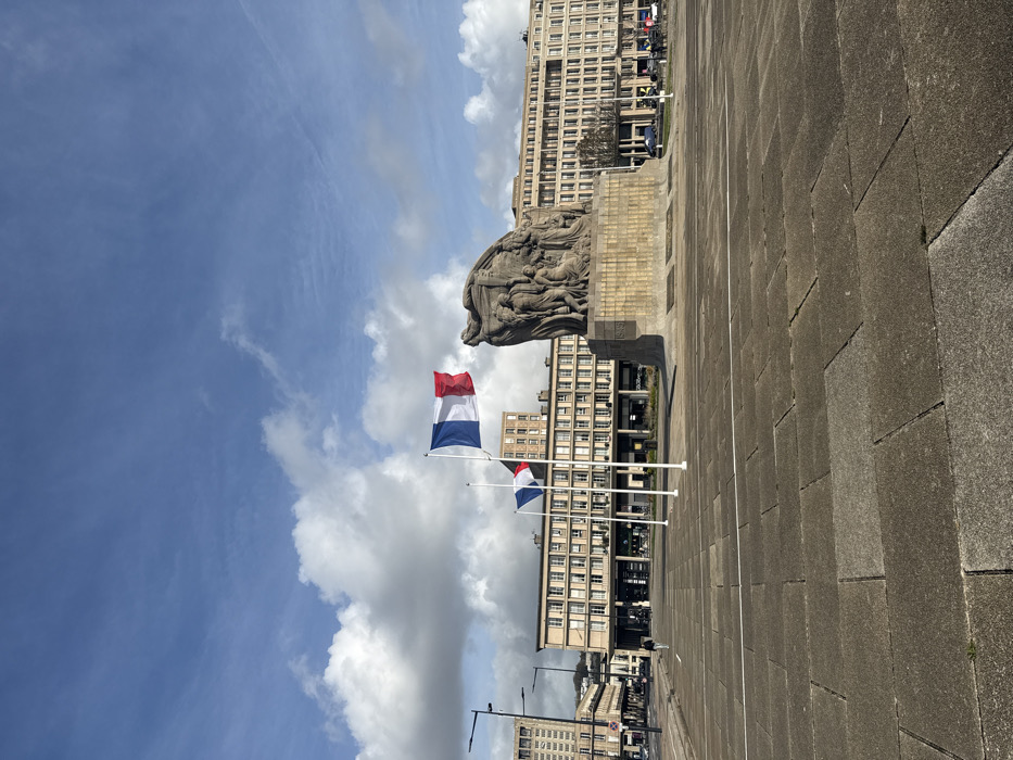 The image depicts the Place de l'Hôtel de Ville in Le Havre, France, under a partially cloudy blue sky with scattered white cumulus clouds. The foreground consists of extensive, light grey rectangular paving stones.
The central subject is the Le Havre War Memorial, a large, monumental stone sculpture situated slightly to the right of the frame's center. It features multiple human figures sculpted in relief on a substantial base, which bears the inscription "1939-1945." To the left of the memorial, three white flagpoles are present. The rightmost flagpole (closest to the memorial) displays a French tricolor flag (blue, white, red vertical stripes) at full mast, while the middle flagpole flies a French tricolor flag at half-mast. The leftmost flagpole is bare.
A single individual, dressed in dark clothing, is observed walking from left to right across the mid-ground of the plaza. In the background, distinctive multi-story modernist buildings, characteristic of Le Havre's post-war reconstruction, line the square. These structures feature light-toned facades with numerous repetitive windows and ground-floor commercial establishments. Several black, multi-armed streetlights are visible on the left side of the scene. Sparse vehicular traffic is present on distant roads bordering the plaza. The scene is illuminated by natural daylight.