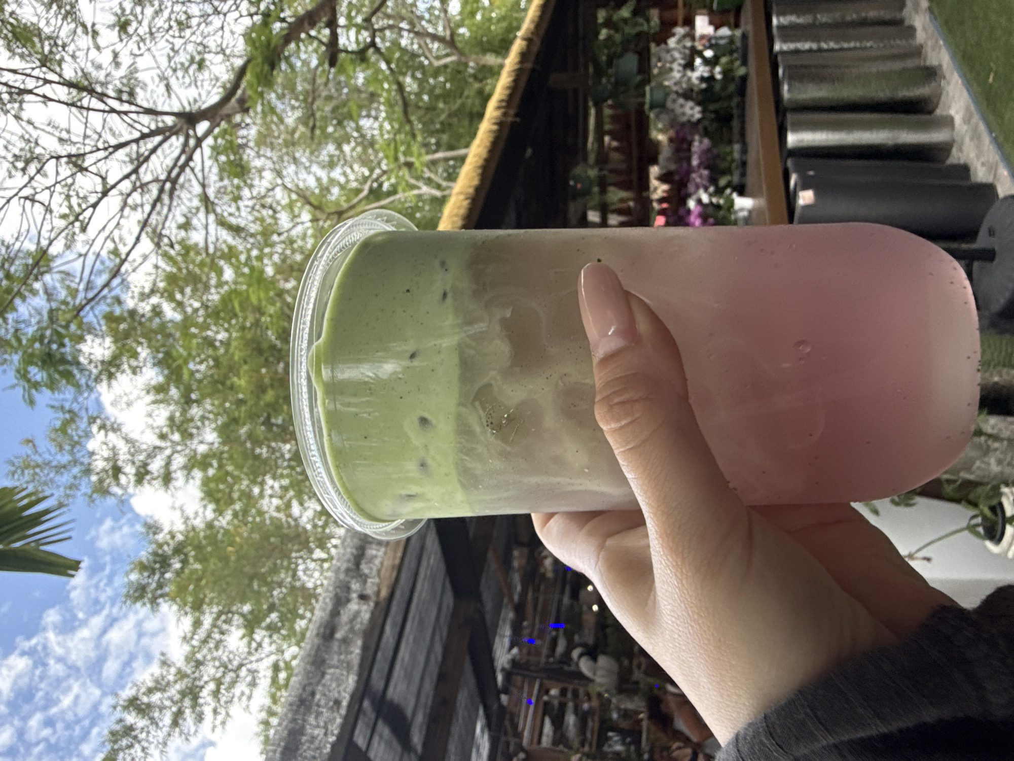 A hand holds a multi-layered beverage in a clear plastic cup. The bottom layer of the drink is pink, the middle is a tan color, and the top layer is green. The hand belongs to a person wearing a gray, long-sleeved shirt. The background includes foliage, building structures, and the sky. The location may be Davie, United States of America.