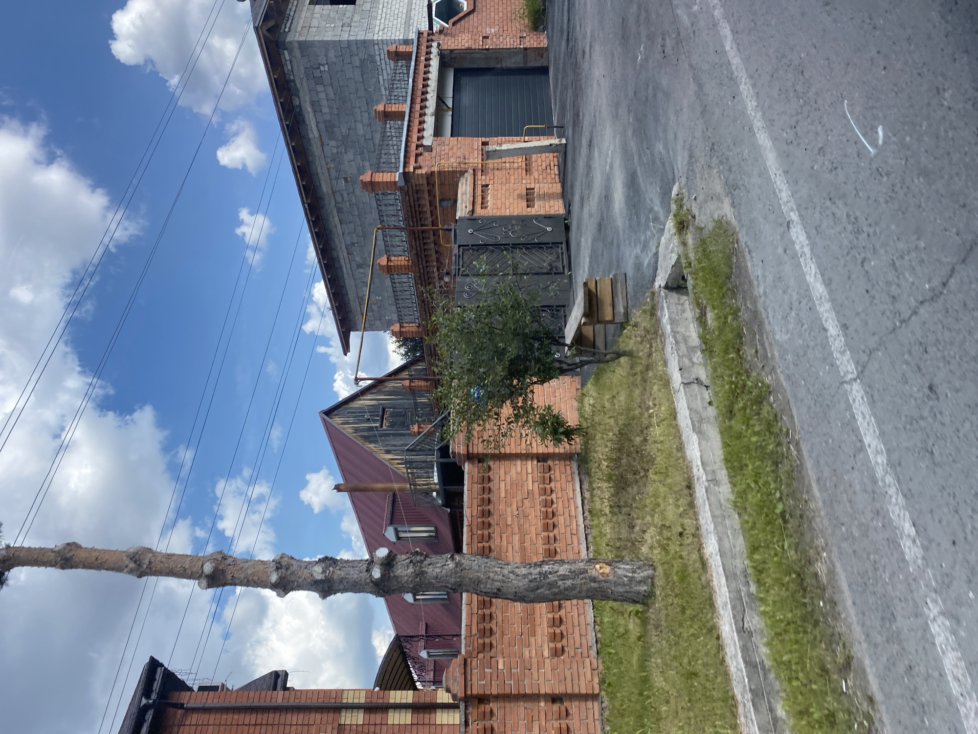 The image depicts a residential area with houses and a fence. In the foreground, a paved road with white markings runs diagonally across the frame. Adjacent to the road is a concrete curb and a patch of grass. A tall, slender tree trunk with a rough texture stands to the left of the frame, its branches pruned.
Behind the tree, a brick wall stretches across the middle ground, separating the road from the property. The wall is made of red bricks and features a decorative pattern with indentations. Beyond the wall, several buildings are visible. To the left, a house with a brick facade and a dark roof is partially obscured. In the center, a building with a red, sloped roof and wooden elements is prominent. To the right, a house under construction, made of grey cinder blocks, dominates the background. This house has a darker grey garage door and a decorative metal railing on an upper level. A metal gate with an ornate design is set into the brick wall in front of the garage. A small, leafy tree grows near the gate. The sky above is blue with scattered white clouds.
The scene is consistent with a residential street in Tyumen, Russia, characterized by a mix of existing and under-construction homes, traditional brickwork, and natural elements.