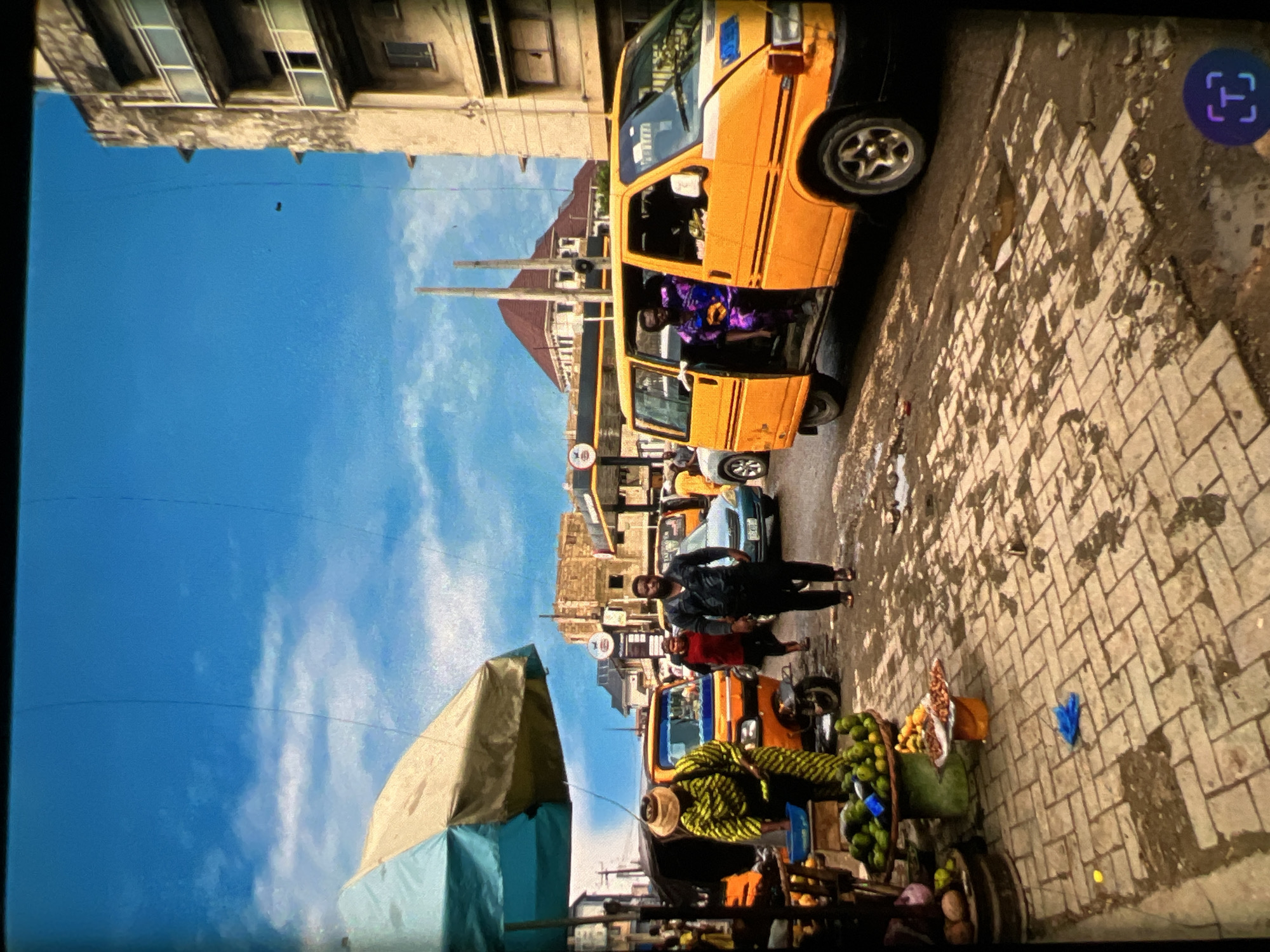 The image depicts a bustling street scene in Lagos, Nigeria, under a bright blue sky with scattered clouds. In the foreground, a market stall displays a variety of fruits, including green mangoes and other produce, with a blue bowl and a woman in a vibrant yellow and green patterned outfit standing nearby. To the right, a yellow commercial van, commonly known as a "danfo," is parked with its doors open, revealing passengers inside. Several people are walking on the cobblestone street, which shows signs of recent rainfall with puddles and wet patches. In the background, more vehicles, market stalls, and multi-story buildings are visible, indicative of a busy urban environment. The architecture and general atmosphere suggest a typical day in a commercial area of Lagos.