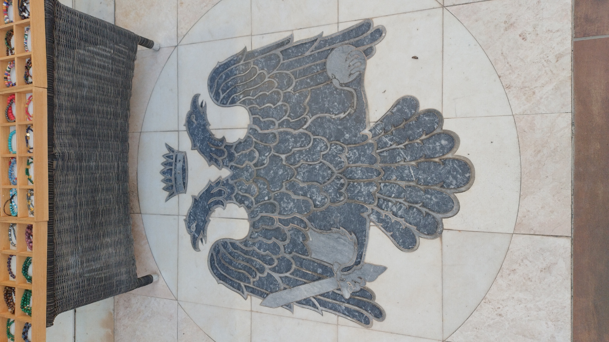 This is a photograph of a decorative stone inlay of an eagle with spread wings, a crown, and a sword. The eagle is depicted in dark gray stone against a lighter, cream-colored tiled background. The inlay is circular, with the eagle positioned in the center. To the left of the inlay is a dark, woven bench or chair. To the far left of the image is a display stand filled with colorful bracelets. The setting appears to be outdoors, possibly a patio or promenade, given the tiling and the presence of outdoor furniture and a retail display. The lighting suggests it is daytime, with no strong shadows indicating a particular time of day, and the weather appears to be clear. The image is a still shot, with no discernable activity taking place. There is no visible text in the image. The overall impression is one of a decorative feature in a public or commercial space.