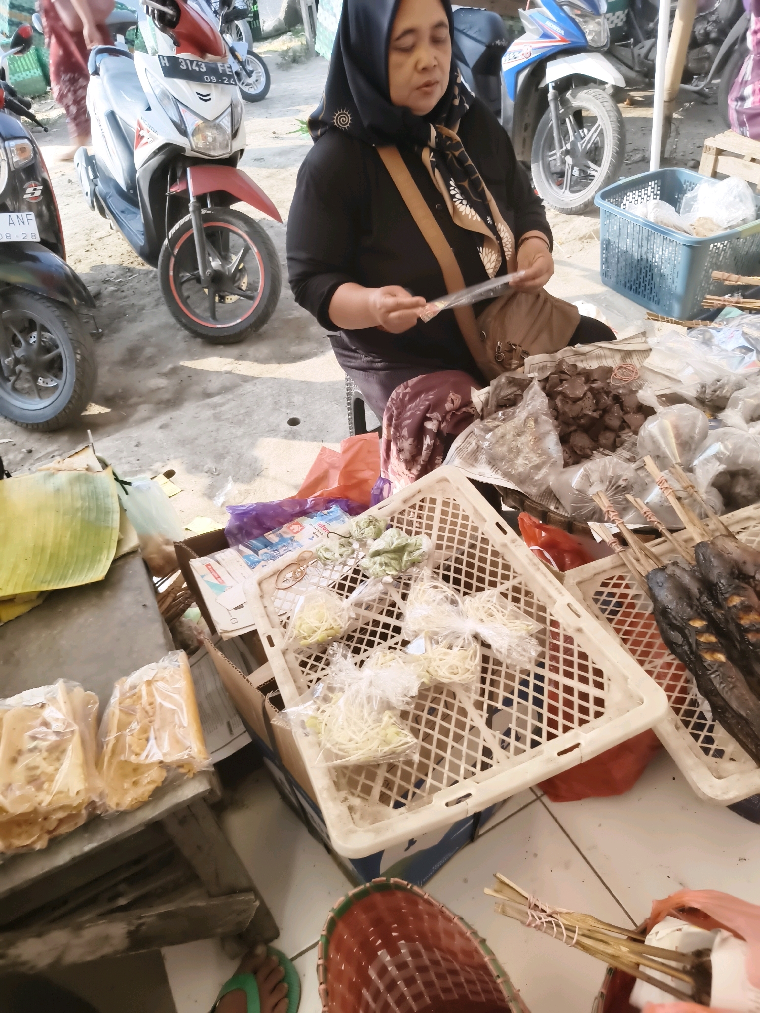 In Guntur, Indonesia, a street market scene unfolds with a woman in a headscarf and black shirt attentively managing her stall. She is seated, and her hands are engaged with a piece of transparent plastic. The stall displays a variety of goods, including what appear to be packaged snacks, a basket filled with raw noodles, and several skewers of grilled fish. Nearby, a blue plastic basket contains more food items. In the background, motorcycles are parked, suggesting a busy area. The ground is a mix of paved and dirt surfaces. The lighting indicates it is daytime.