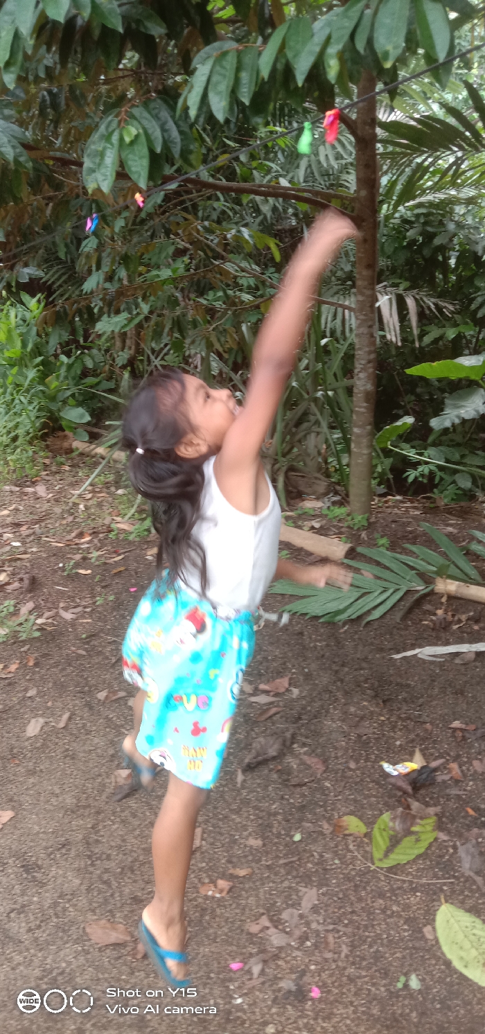 A young girl in Ranugedang, Indonesia, is depicted outdoors, captured mid-action while jumping or stretching upwards. She wears a white sleeveless top, a light blue patterned skirt adorned with cartoon characters and text, and blue flip-flop sandals. Her dark hair is pulled back in a low ponytail. Her right arm is extended vertically and appears blurred from motion, reaching towards a black string stretched horizontally across the upper-mid portion of the frame. Attached to this string are several small, brightly colored items, including pink, green, orange, and blue objects, possibly balloons or decorations. Her head is tilted back, and her mouth is open as she looks upwards towards these items. The background consists of dense green foliage from various broad-leafed trees and palm-like plants. The ground is exposed brown soil, sparsely covered with scattered dry leaves, twigs, and a few small pink flower petals. A discarded wrapper and a large green leaf are visible on the ground.