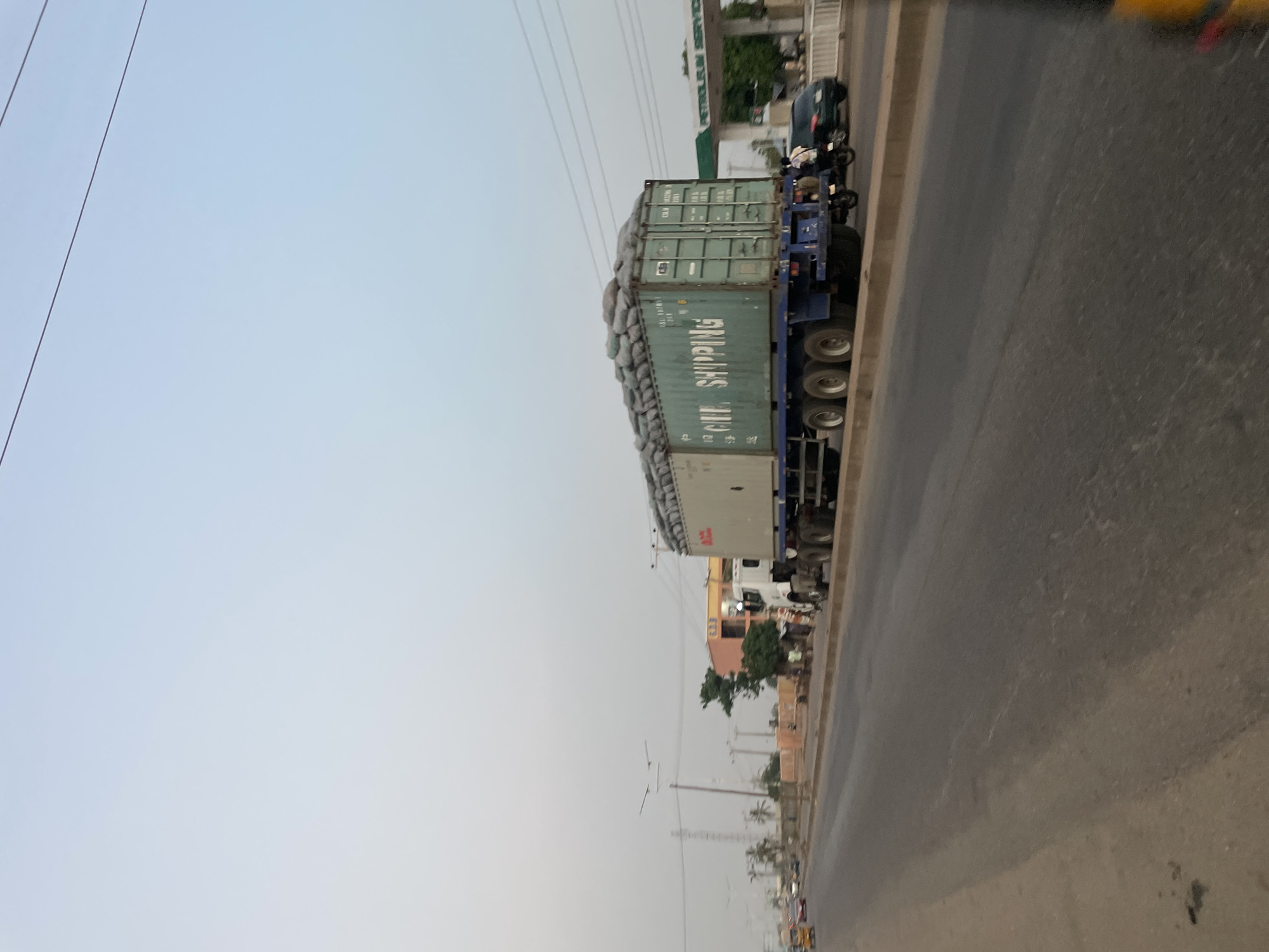 A slightly tilted, medium-shot captures a commercial truck stopped on a paved road in what appears to be Hayin Banki, Nigeria, based on visual characteristics of the built environment. The truck is carrying two stacked, green cargo containers filled with what appear to be bagged goods, stacked high over the top of the shipping containers. The bags on top of the containers appear to be covered with light-colored fabric. A motorcycle is beside the truck with two riders. Buildings, vegetation, and power lines are visible along the roadside. The sky is overcast and the environment shows potential signs of atmospheric haze or dust. The road surface is worn and shows signs of use. There are a number of buildings and signs of commercial activity nearby.