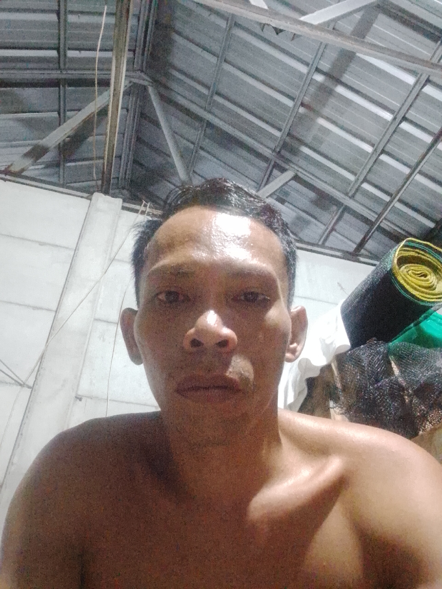 The image is a selfie of a shirtless adult male with dark hair. The man appears sweaty and is looking directly at the camera. He is in an indoor setting with a corrugated metal roof above him. To the left of the frame is a white wall with visible wiring. To the right, there are stacked objects including a rolled yellow and green object and black mesh. The location is in an unspecified indoor setting, Bukit Ketok, Indonesia.