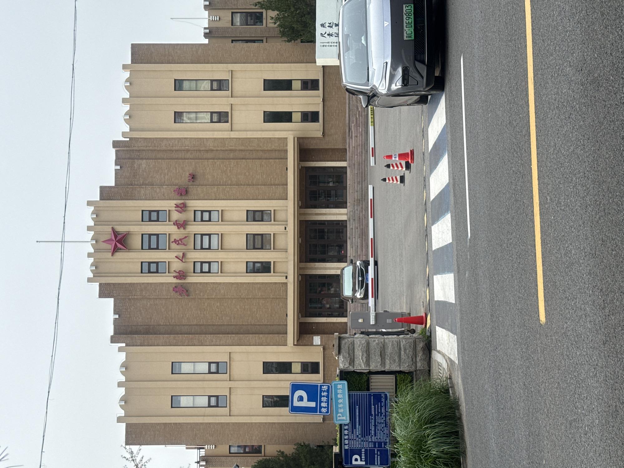 The image captures the exterior of a building with distinct architectural features, identified by signage as the "Shanhaiguan District Cultural Center" in Qinhuangdao, China. The building is constructed of light brown brick and tan concrete, with a prominent red star emblem above the entrance. Tall, narrow windows are symmetrically placed across the facade.
In front of the building, a paved area serves as a driveway leading to a white barrier gate. Two vehicles are visible: a dark-colored sedan stopped at the gate and a modern, light gray electric vehicle in the foreground, partially obscuring a pedestrian crossing. Several orange traffic cones are strategically placed on the pavement.
To the left of the entrance, a stone pillar supports multiple blue and white parking signs, indicating a paid parking area. The overall scene suggests a functional civic building within an urban setting, under an overcast sky.