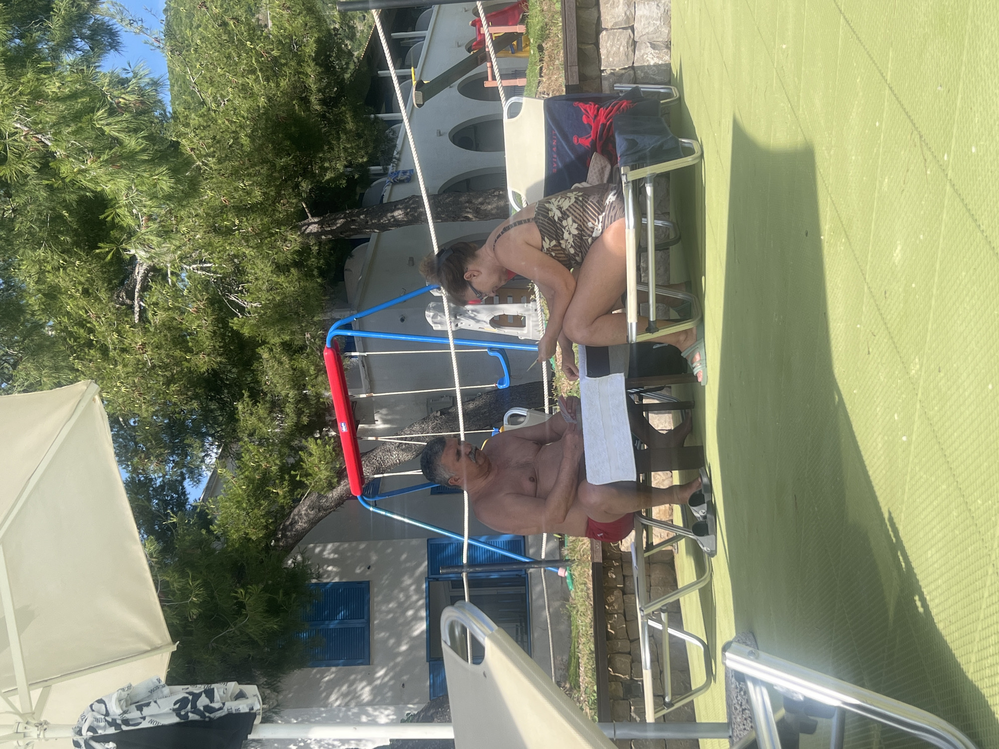 This image captures an outdoor scene in Budva, Montenegro, on a bright, sunny day. Two adults, a man and a woman, are seated opposite each other at a small, light-colored rectangular table, engaged in a card game.
The man, positioned on the left, is shirtless and wears red swimming shorts and grey slide sandals. He has a mustache and is holding a fanned deck of cards in his hands. The woman, on the right, is wearing a patterned one-piece swimsuit, sunglasses, and light green slide sandals. She is looking down at an object in her hands, possibly also cards or a small device. A blue towel with a red crown design and "SVILANIT" text is draped over the back of her chair. Both individuals are sitting on lightweight folding chairs with metal frames and light-colored fabric.
The ground beneath them is covered with a light green, textured matting, likely for outdoor recreational use. In the foreground on the left, part of another sun lounger is visible. Above and to the left, a cream-colored beach umbrella partially obstructs the view, with a black t-shirt and a patterned t-shirt hanging from its pole.
In the background, a children's play area features a red and blue swing set. Behind the swing set and the man, a white building with blue louvered shutters and dark windows is visible. Further to the right and back, another white building with several arched openings and a tiled roof is partially obscured by lush green trees, which dominate the background landscape. A stone wall section runs behind the woman. White ropes are strung horizontally across the middle ground. The clear blue sky is visible above the tree canopy and distant hills or mountains. Strong sunlight casts shadows on the green matting.