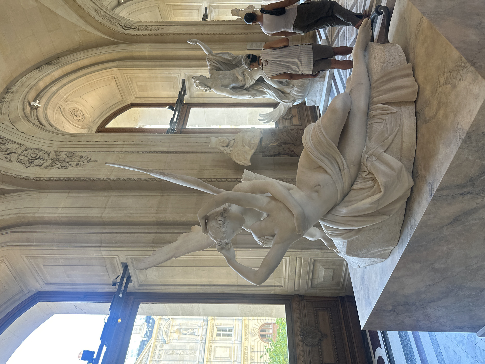 The image depicts the interior of a grand museum, likely the Louvre Museum in Paris, France, given the architectural style and the presence of classical sculptures. In the foreground, a prominent marble sculpture of Cupid and Psyche is displayed on a marble pedestal. The sculpture portrays Cupid embracing Psyche, who is depicted lying down with her arms around him. Behind this central sculpture, other marble statues and busts are visible, including a large statue of a male figure with an outstretched arm. Two individuals, a man wearing a striped tank top and shorts, and a woman in a white tank top and dark pants, are walking away from the camera in the mid-ground. The museum's interior features high arched ceilings, large windows offering a view of external architecture, and intricately patterned flooring. The lighting suggests a daytime scene, with natural light streaming in from the windows.