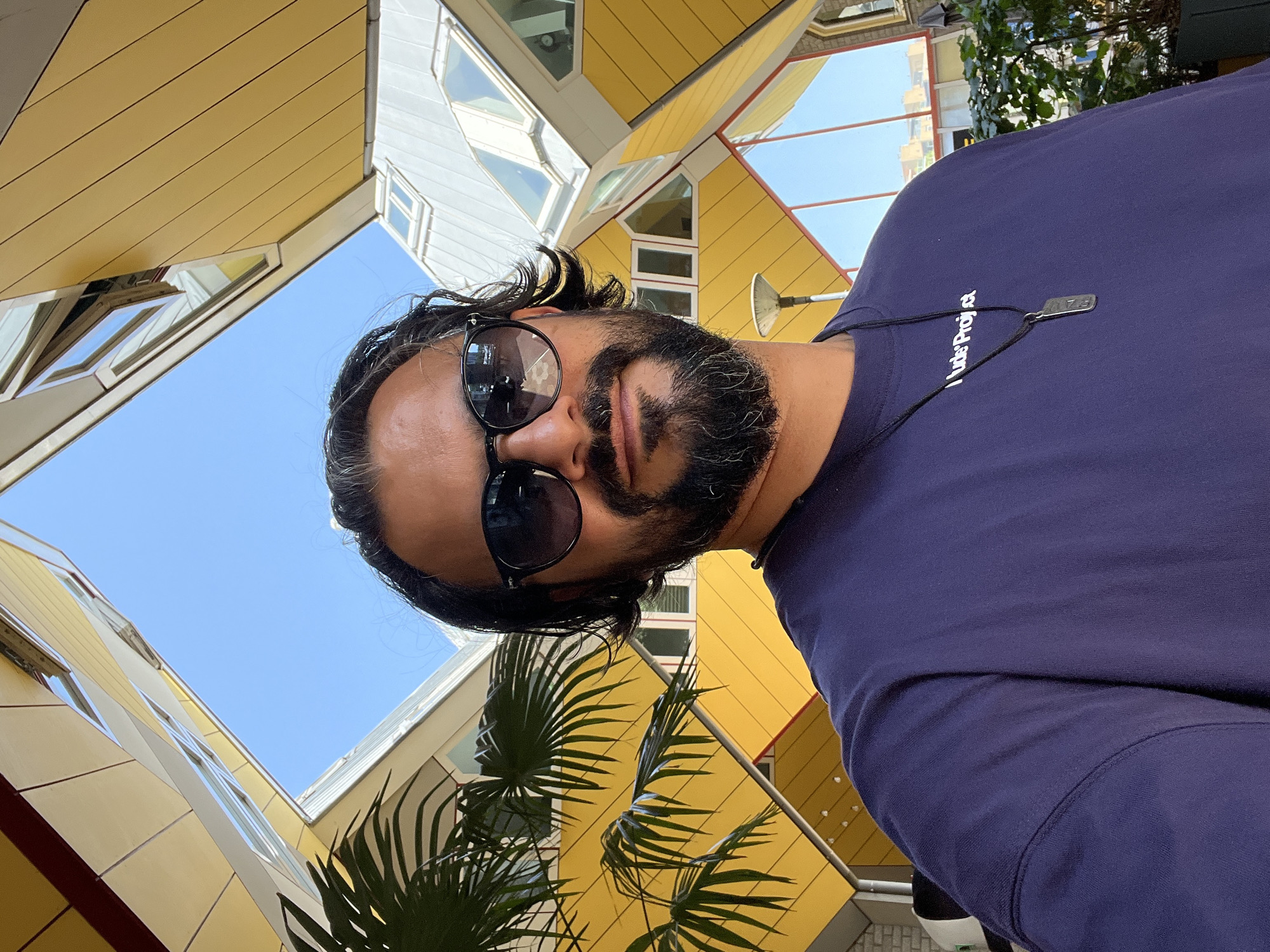 A man with dark hair and a beard is taking a selfie in front of the Cube Houses in Rotterdam, Netherlands. He is wearing sunglasses and a dark blue t-shirt with a white inscription and a pendant necklace. The Cube Houses, with their distinctive yellow and white angular facades, are visible in the background, along with a clear blue sky. A palm tree is partially visible on the left side of the image.