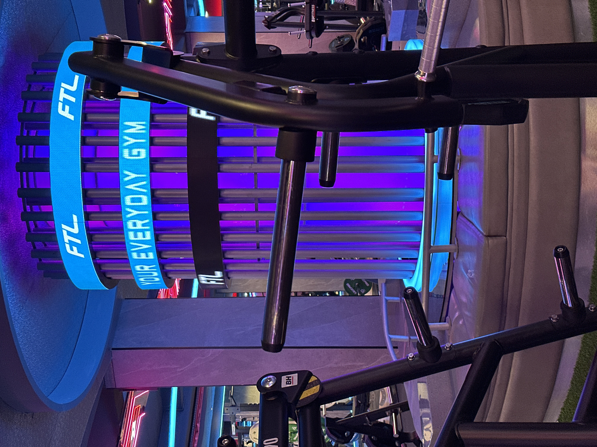 The image shows fitness equipment, specifically a Lat Pulldown machine, in what appears to be a modern gym with neon lighting. The machine has a blue cylindrical element with white text reading "FTL YOUR EVERYDAY GYM". The gym appears well-equipped with other exercise machines visible in the background. Based on the text on the machine, it is likely located at FTL GYM in North Kuta, Indonesia.