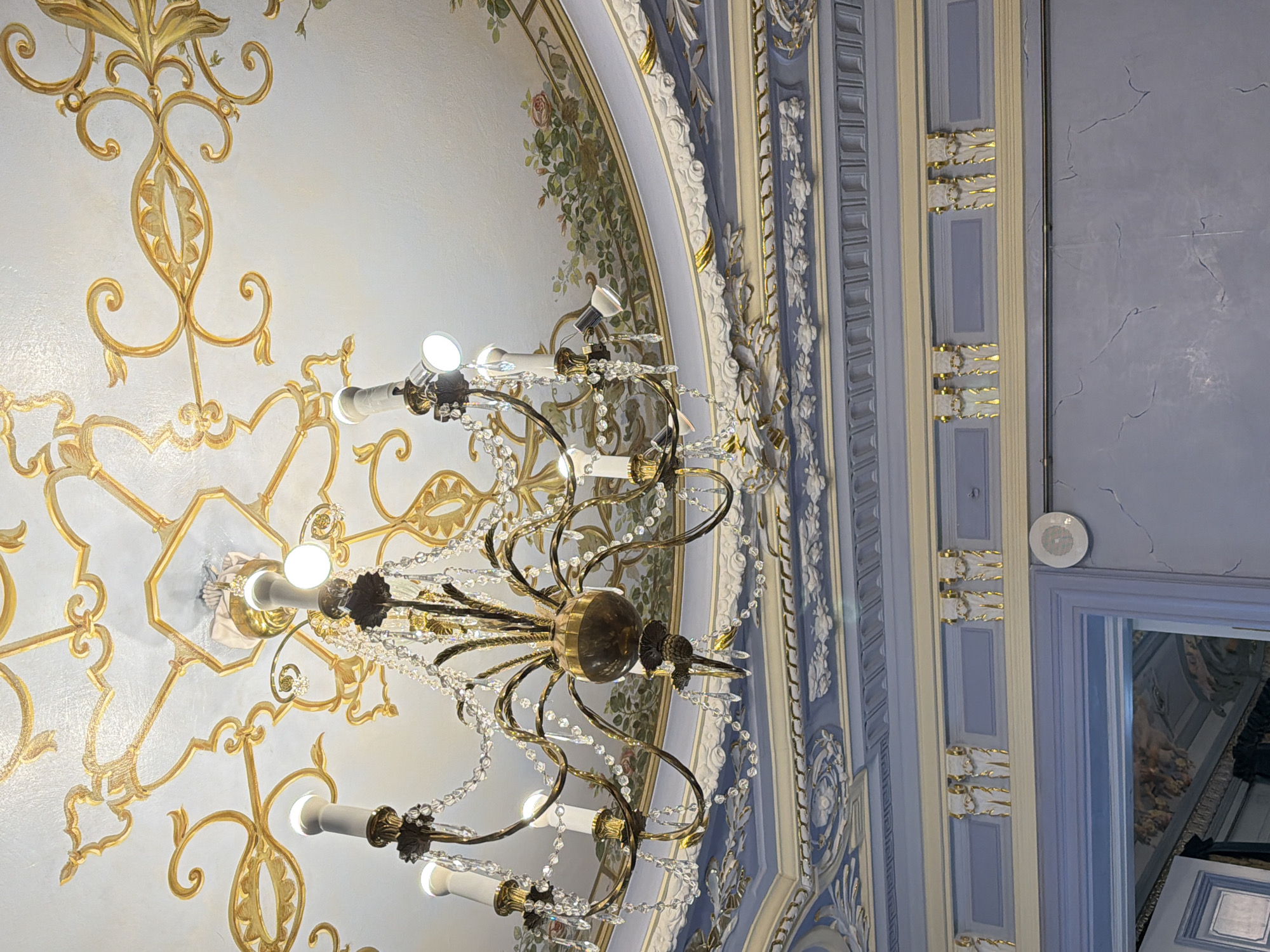 The image displays an ornate, classic interior with a gilded ceiling adorned with intricate scrollwork. A crystal chandelier with multiple lights hangs from the center of the ceiling. Below the ceiling, there is a decorative frieze with floral motifs. The walls are painted a pale blue with a subtle crackled effect and feature a horizontal band of decorative gold elements. A doorway with a dark interior is visible at the bottom left. The scene is well-lit, likely from the chandelier and possibly ambient room lighting. The overall impression is one of opulence and historical grandeur. This photograph was taken in Nizhny Novgorod, Russia.