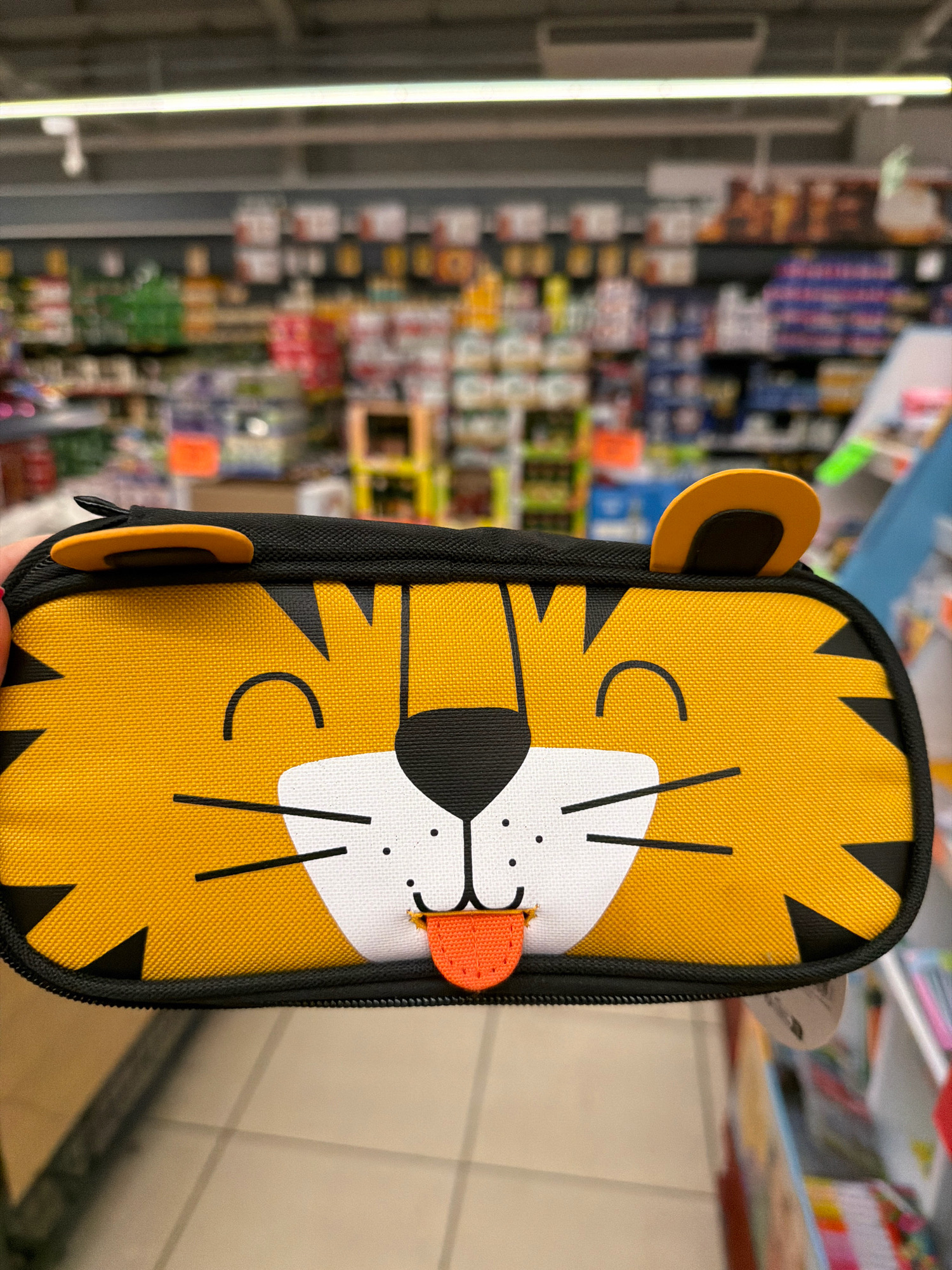 The image shows a close-up view of a pencil case designed to look like a tiger's face, held in a person's hand in what appears to be a supermarket aisle. The pencil case is yellow with black tiger stripes, black ears, and a white face with a black nose, closed eyes indicated by curved lines, and a small pink tongue sticking out. The background is blurred but shows shelves stocked with various products, typical of a retail environment. Based on the visual cues and the context of a supermarket, this scene could be located in a commercial area. Without further information or context, it is impossible to definitively place this specific scene in Wrocław, Poland.