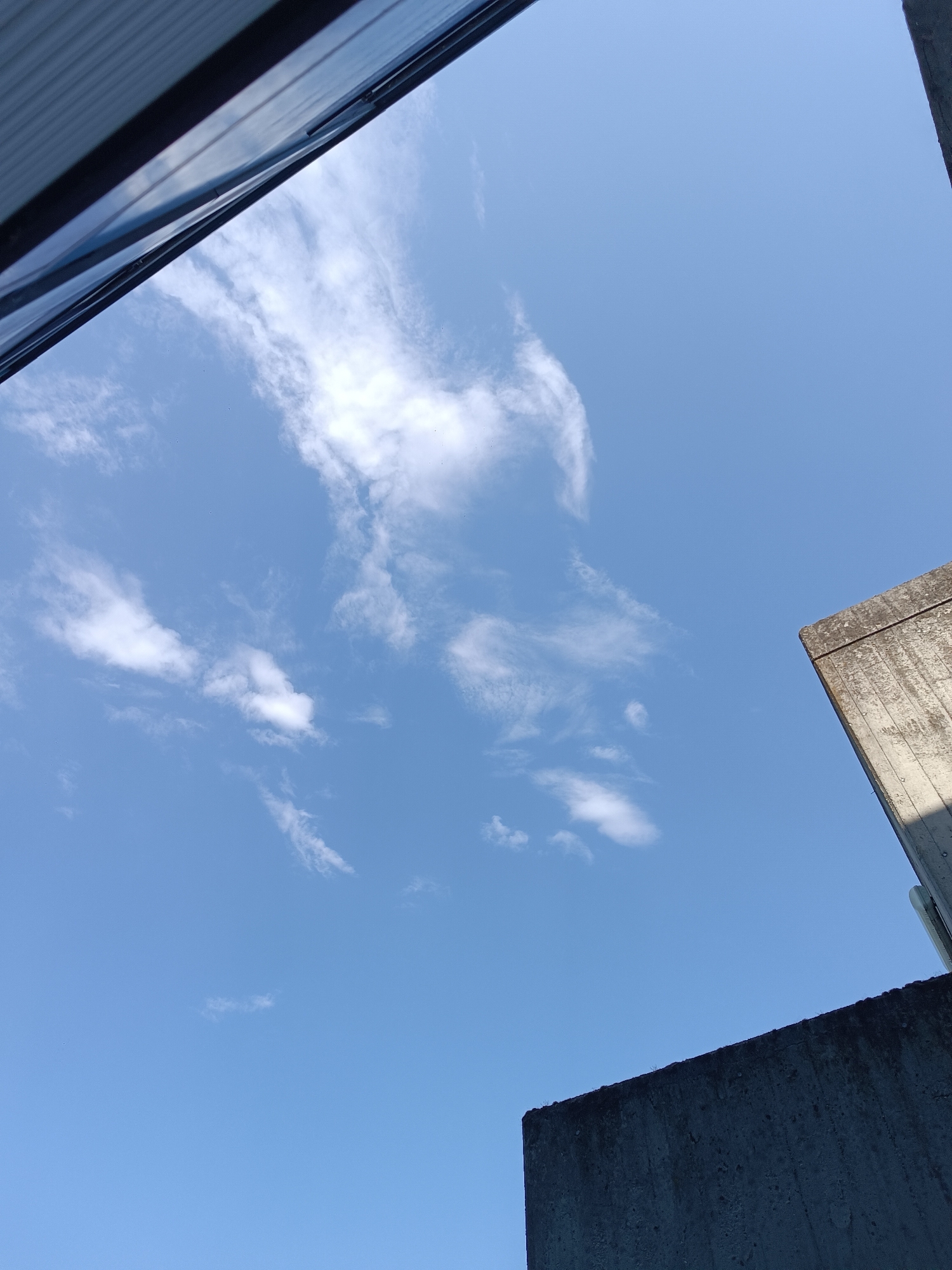 The image shows a clear blue sky with scattered wispy clouds, viewed from an upward angle. The scene is framed by two concrete structures and a portion of what appears to be a modern building with a ribbed metal roof or siding. In the lower right and upper right corners, sections of rough, grey concrete walls are visible, suggesting an urban or industrial setting. The perspective is that of looking up from ground level between buildings. There are no people or animals visible in the image. The lighting suggests it is daytime, with the sun casting shadows on the concrete surfaces. The location is identified as Casalmaggiore, Italy.