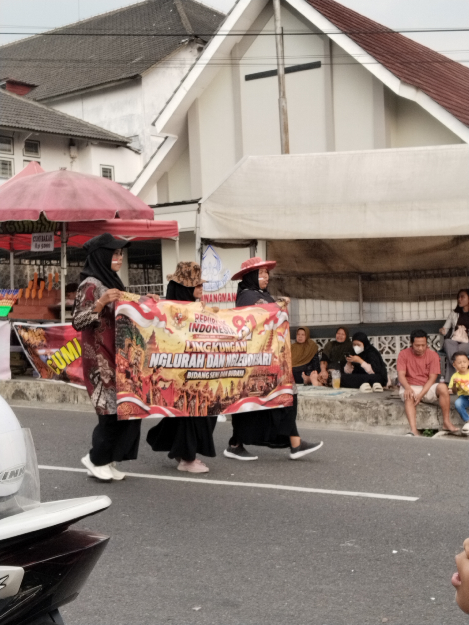 This is a photograph taken in Tawangmangu, Indonesia. In the foreground, a motorcycle is partially visible. A procession is underway on a paved road. Two women, dressed in traditional Indonesian attire and headscarves, are walking from left to right, holding a large banner. The banner is colorful and features images of traditional Indonesian culture, including what appear to be dancers and temples. Text on the banner is in Indonesian.
Behind the procession, spectators are seated on a raised concrete wall. They are casually dressed, and some are wearing masks. A red umbrella is visible on the left side of the image, suggesting a market or vendor area. In the background, a building with a distinctive white facade and a red-tiled roof stands under a cloudy sky. The architecture of the building, particularly its gabled roof and angled white walls, suggests it might be a place of worship or a public building. The overall scene depicts a local cultural event or celebration in Tawangmangu.