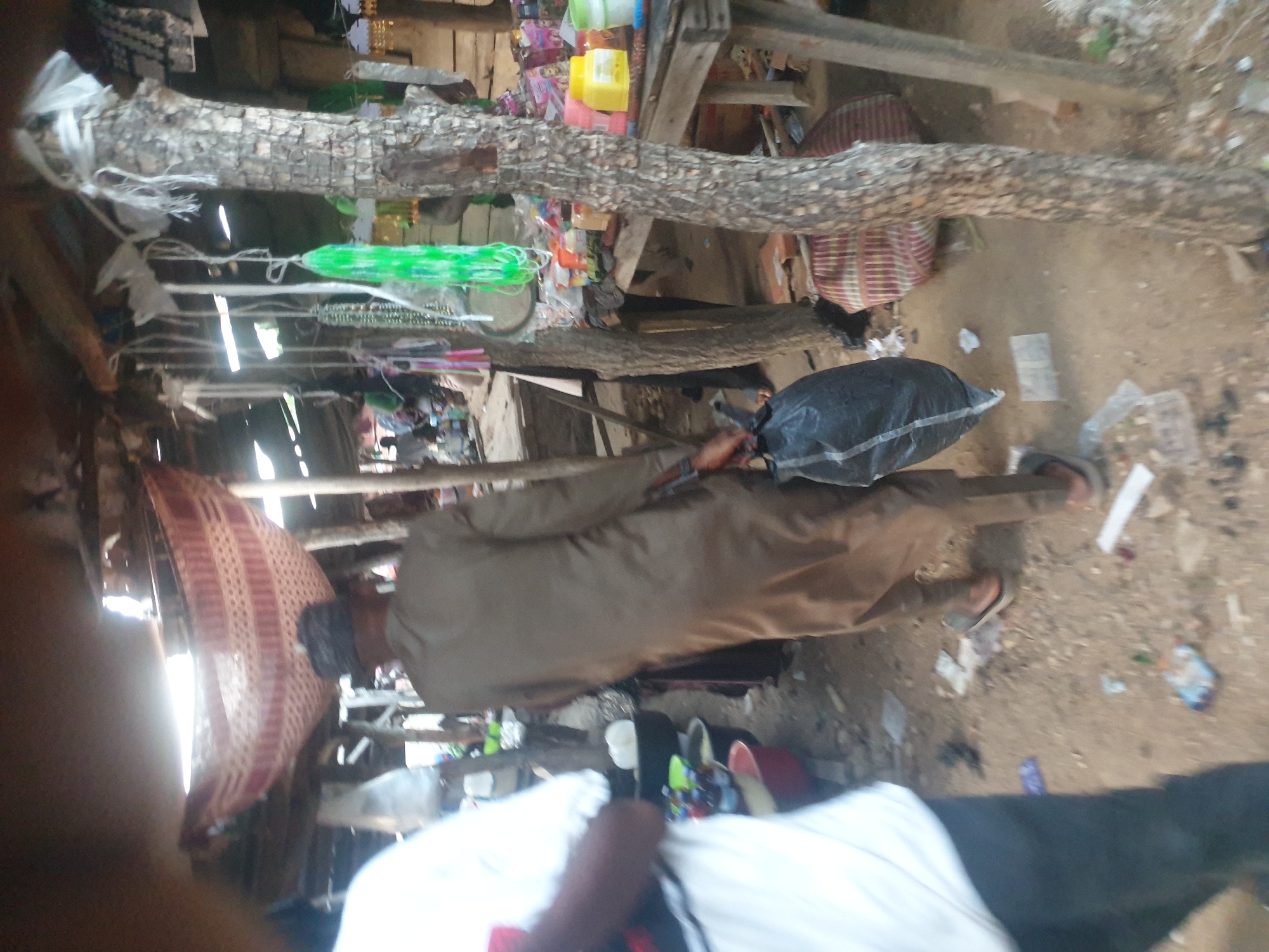 The image shows a cluttered outdoor space in Unknown, Unknown. A person in a brown robe is seen holding a black plastic bag. A weathered wooden structure runs horizontally across the upper portion of the image. An object resembling a mat or folded material is on the left, and debris litters the ground.
