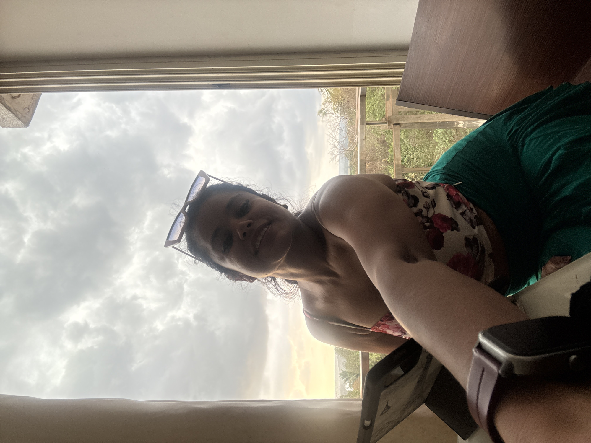 A woman is taking a selfie indoors in Sangolda, India. She is wearing sunglasses on her head, a floral print top, and green pants. Her left arm, which is wearing a smartwatch, is extended towards the camera.
Behind her is a window with a view of a cloudy sky, trees, and buildings. A wooden cabinet is visible to her right. The light suggests it could be late afternoon or early evening.