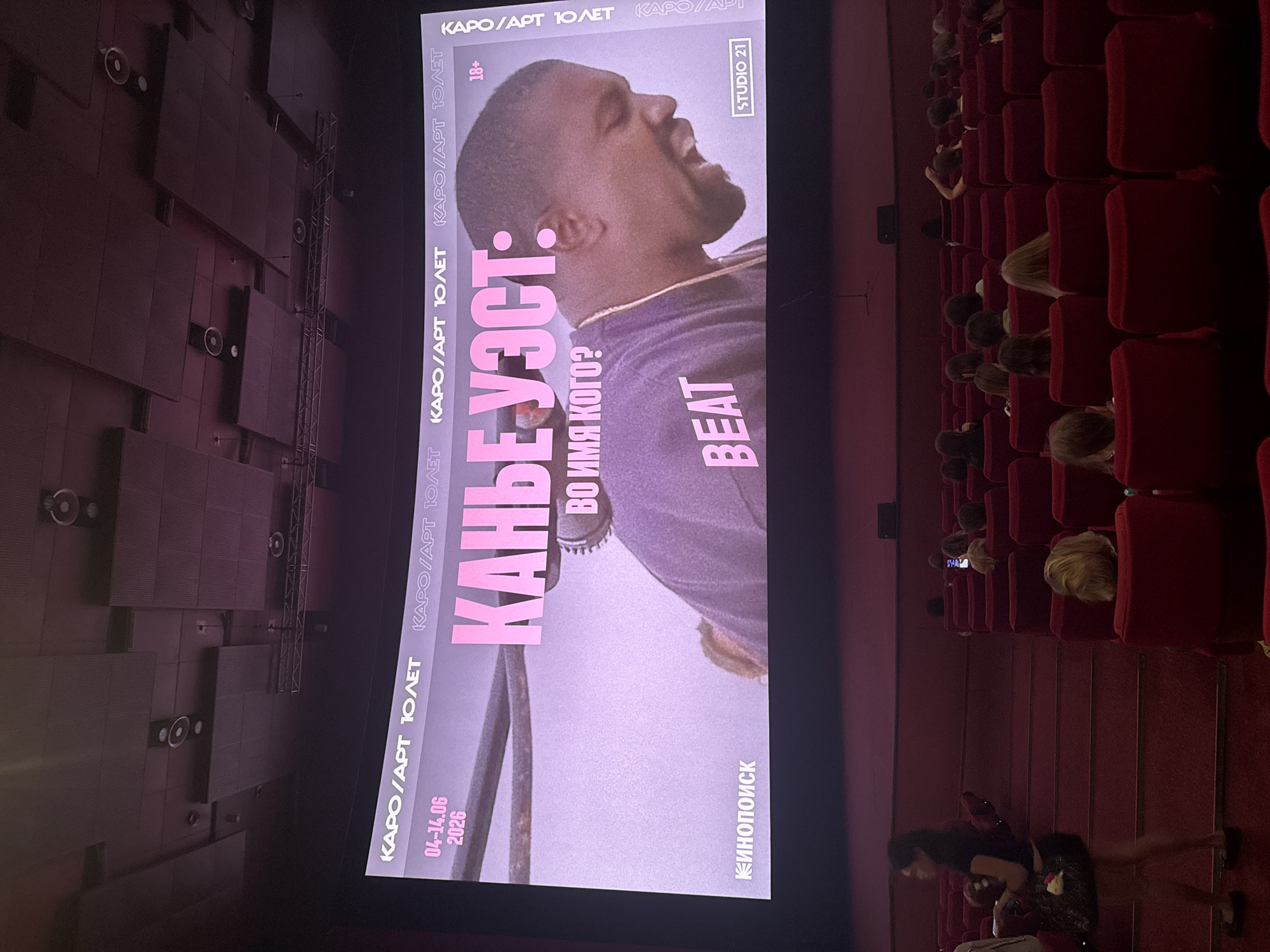 In a cinema in Moscow, Russia, a movie poster is displayed on a large screen. The poster features a close-up of Kanye West, with the words "Kanye West" and "Beat" in large pink letters. Below "Kanye West" is the text "ВО ИМЯ КОГО?" which translates to "In whose name?". To the left of the image, dates "04-14.06 2026" are visible, along with the text "КИНОПОИСК" which is a Russian film database. The top of the screen displays "KARO/ART 10 ЛЕТ" which translates to "Karo/Art 10 Years". The audience is seated in red cinema seats, facing the screen. A woman is standing on the left side of the frame, near the stairs leading to the seats. The overall lighting is dim, with the screen being the primary light source.