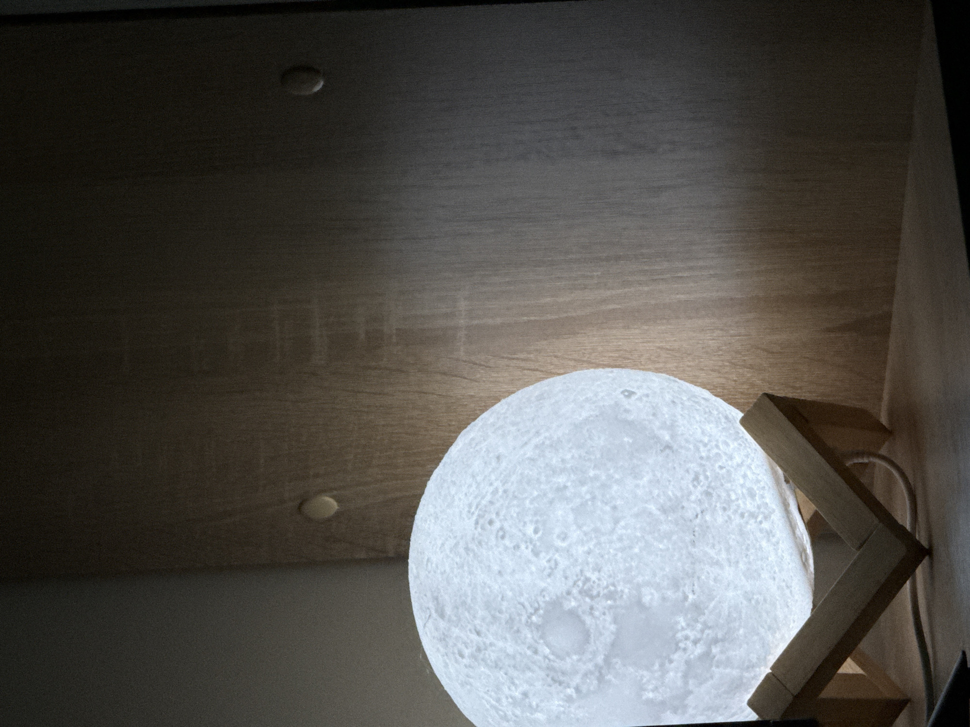 The image shows a close-up of a round, white object that resembles the moon, likely a lamp, resting on a wooden stand. The lamp is illuminated, casting a soft glow on its textured surface, which is designed to mimic lunar craters. The wooden stand is made of light-colored wood and has a zig-zag or folded design. In the background, a wooden surface, possibly a shelf or ceiling, is visible with subtle wood grain. There are two small, round indentations or fixtures on this wooden surface. The overall lighting is dim, suggesting an indoor setting, perhaps in the evening or with ambient light. The focus is on the moon lamp and its immediate surroundings.