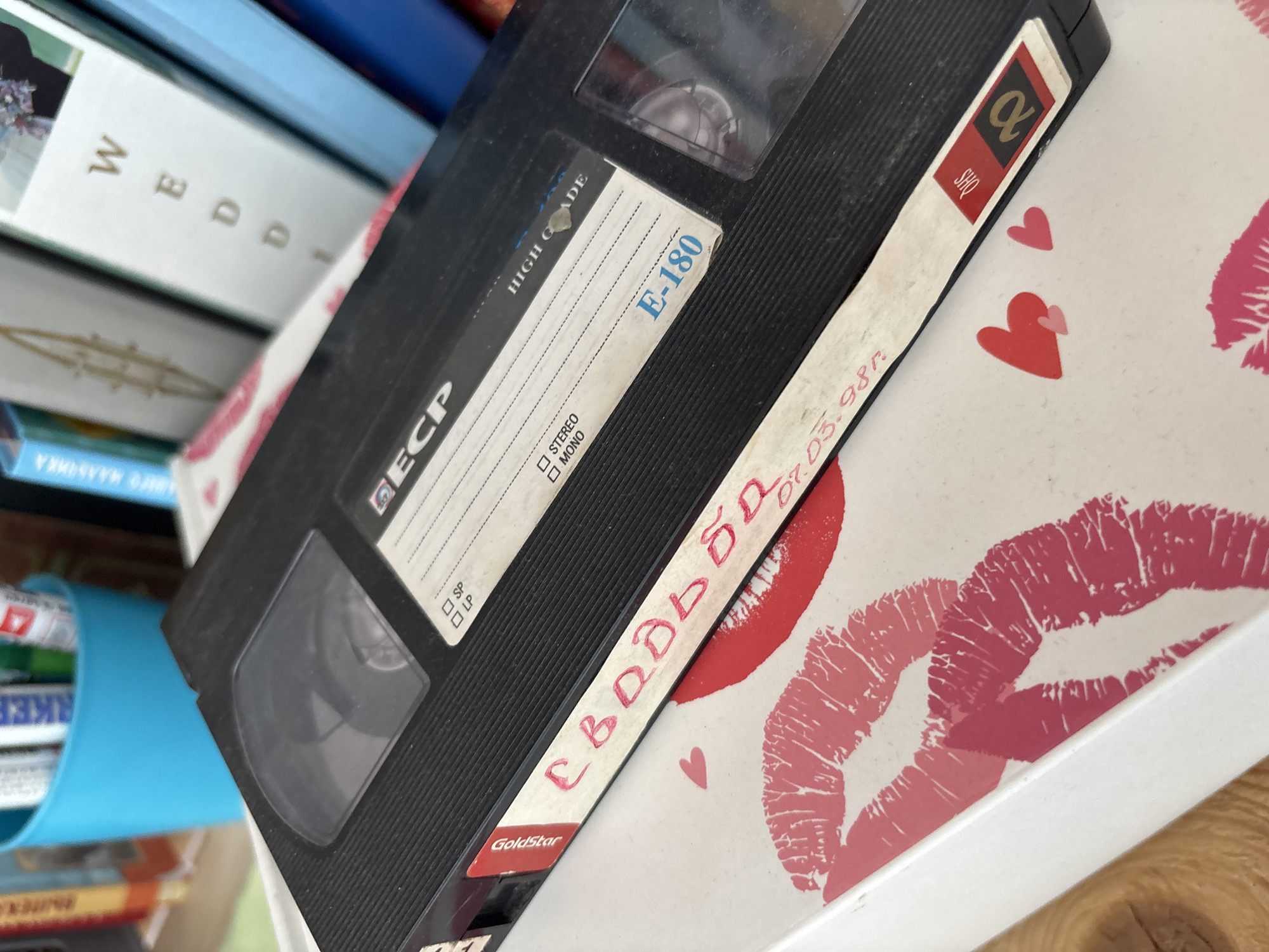 The image displays a close-up of a VHS tape resting on a white surface decorated with pink and red lipstick kisses. The VHS tape is black with a clear window showing the reels. A white label on the tape has handwritten text in red and some printed text. The handwritten text appears to be in Cyrillic script and reads "Свадьба" which translates to "Wedding". Below this, there is a date written as "07.03.98r", likely indicating March 7, 1998. Printed text on the tape includes "GoldStar" and "ECP". In the background, blurred objects are visible, including a blue container holding pens or markers and books with titles partially obscured. The background is out of focus, making it impossible to determine the location within the city of Sochi, Russia or any other specific details about the environment. There are no people or discernible actions depicted in the image.