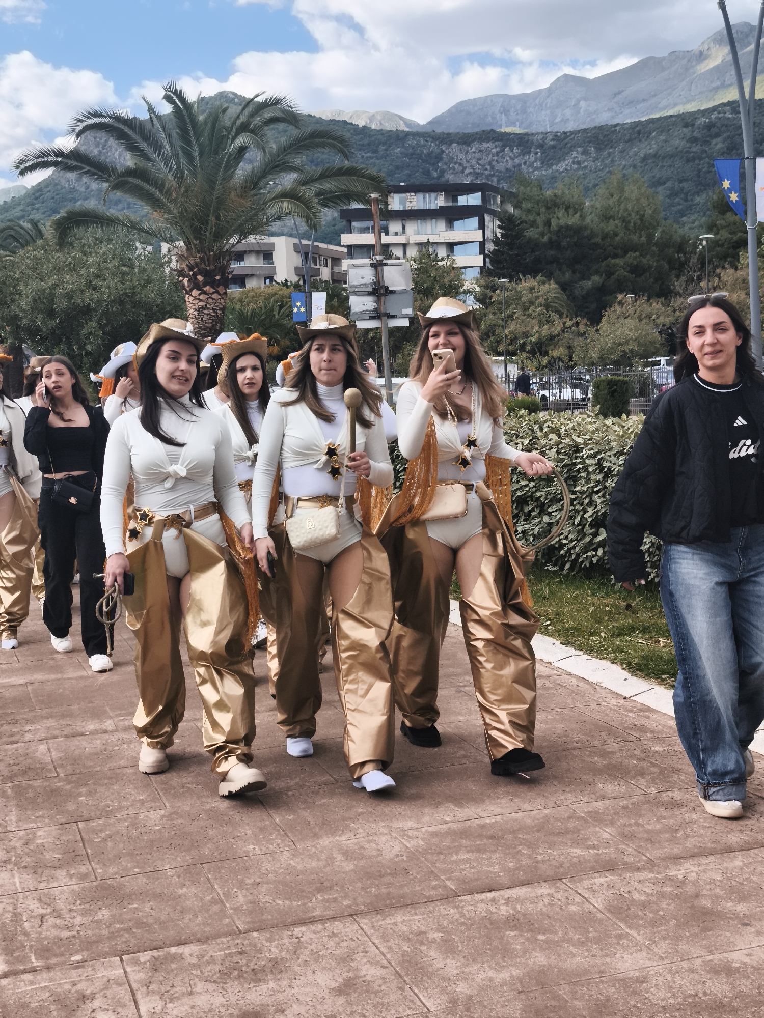 The image shows a group of young women dressed in white tops, gold chaps, and cowboy hats walking along a paved walkway. One woman is walking in the opposite direction in jeans and a black jacket. The background includes a palm tree, a building, mountains, and trees. A flagpole with the flag of Europe is visible. The location is a street or public space.