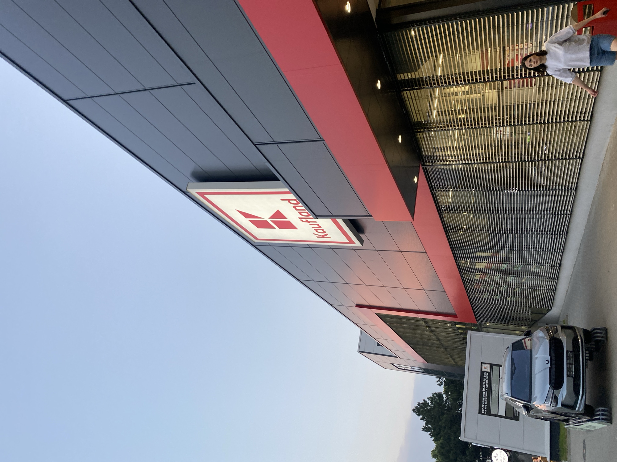 The image shows the exterior of a Kaufland supermarket in Chișinău, Moldova. The building has a modern architectural design with a dark grey facade and a prominent red band. A large Kaufland logo with a red "K" is visible on the upper part of the building. In front of the store, a silver Skoda Kamiq is displayed on a platform with rollers, suggesting it might be part of an advertisement or promotion. A woman in a light-colored shirt and denim shorts is walking in front of the store's large, slatted windows. The sky is a clear light blue, indicating it is daytime, likely late afternoon or early evening due to the lighting. The environment appears to be an outdoor commercial area.