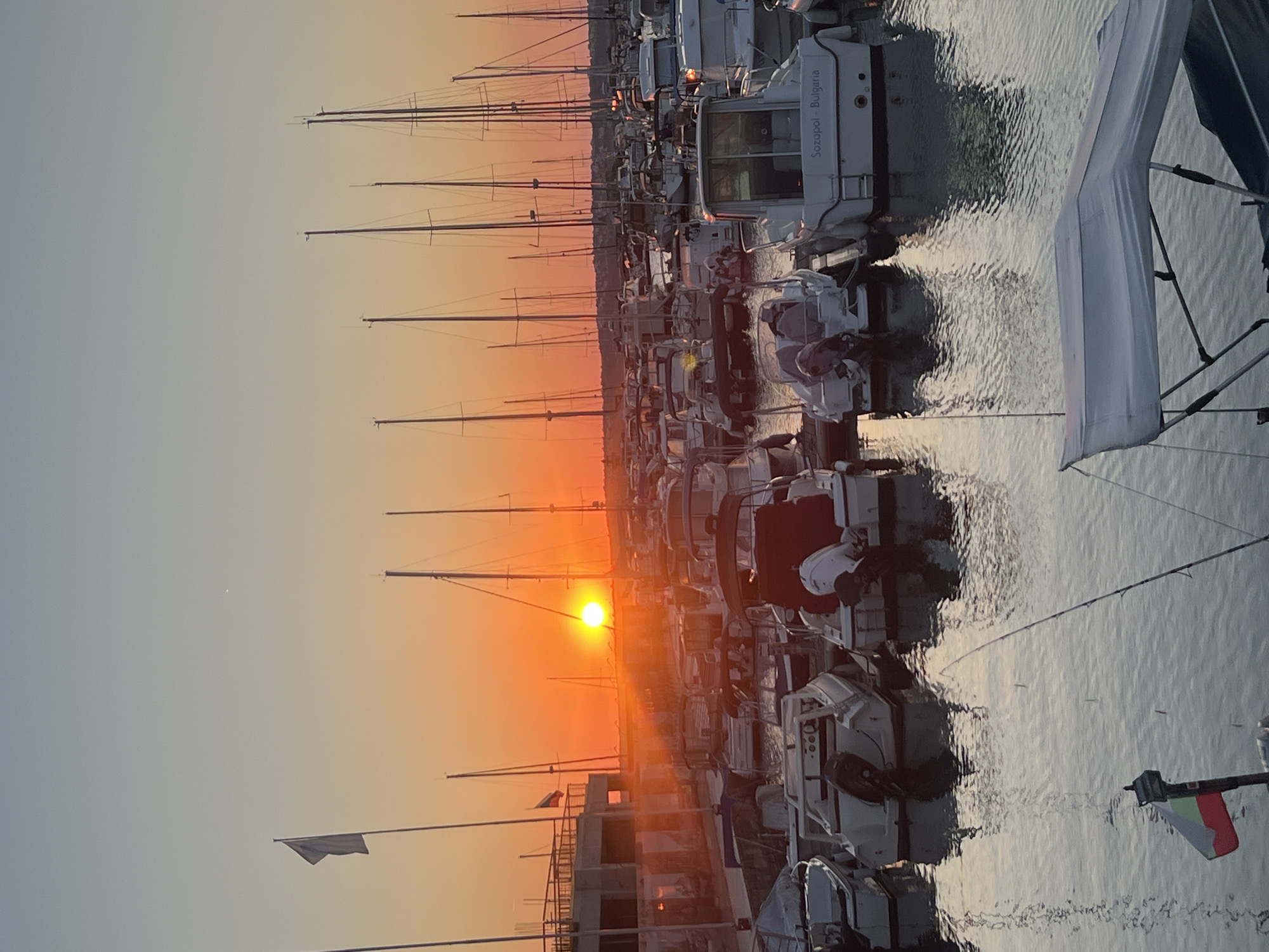 The image depicts a harbor scene in Sozopol, Bulgaria, during sunset. The sun, a bright orb, is low on the horizon, casting a warm orange glow across the sky and water. Numerous sailboats and motorboats are docked in the harbor, their masts creating a vertical pattern against the twilight sky. Some of the boats have their covers on, while others appear ready for sailing. The water is calm, reflecting the colors of the sunset and the silhouettes of the boats. A flag is visible on a pole on one of the boats, displaying the colors red and green on a white background, representing the Bulgarian flag. On the shore in the background, a modern building structure is visible, possibly a marina building or a restaurant, with some construction elements suggesting ongoing development. A small white speck, possibly a distant boat or a light fixture, is visible in the upper right portion of the sky. The overall atmosphere is serene and picturesque, characteristic of a coastal town at dusk.