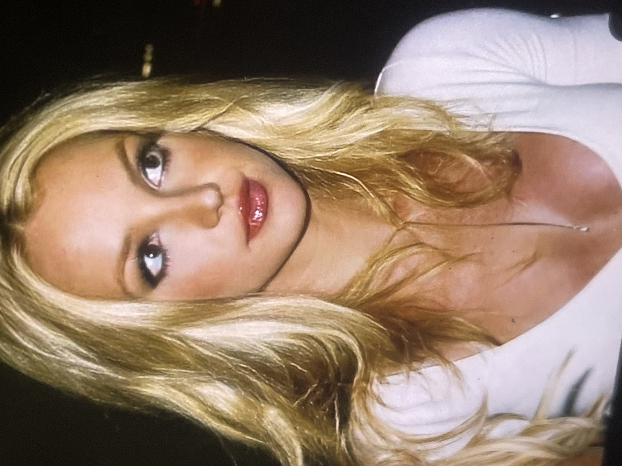 A close-up portrait features a young woman with long, wavy, golden-blonde hair framing her face. Her head is slightly tilted, and she looks directly at the viewer with light-colored eyes defined by dark eyeliner and smoky eyeshadow. Her lips are full and glossy, colored with a vibrant red or deep pink lipstick. She wears a form-fitting, light-colored, long-sleeved top, possibly white, with a low neckline, and a delicate necklace with a small pendant is visible. The background is dark and out of focus, showing indistinct blurry lights, suggesting an indoor setting or a nighttime environment. The image appears to be a posed shot, capturing a moment of glamour, potentially during an evening event in Poltava, Ukraine. There is no visible text.