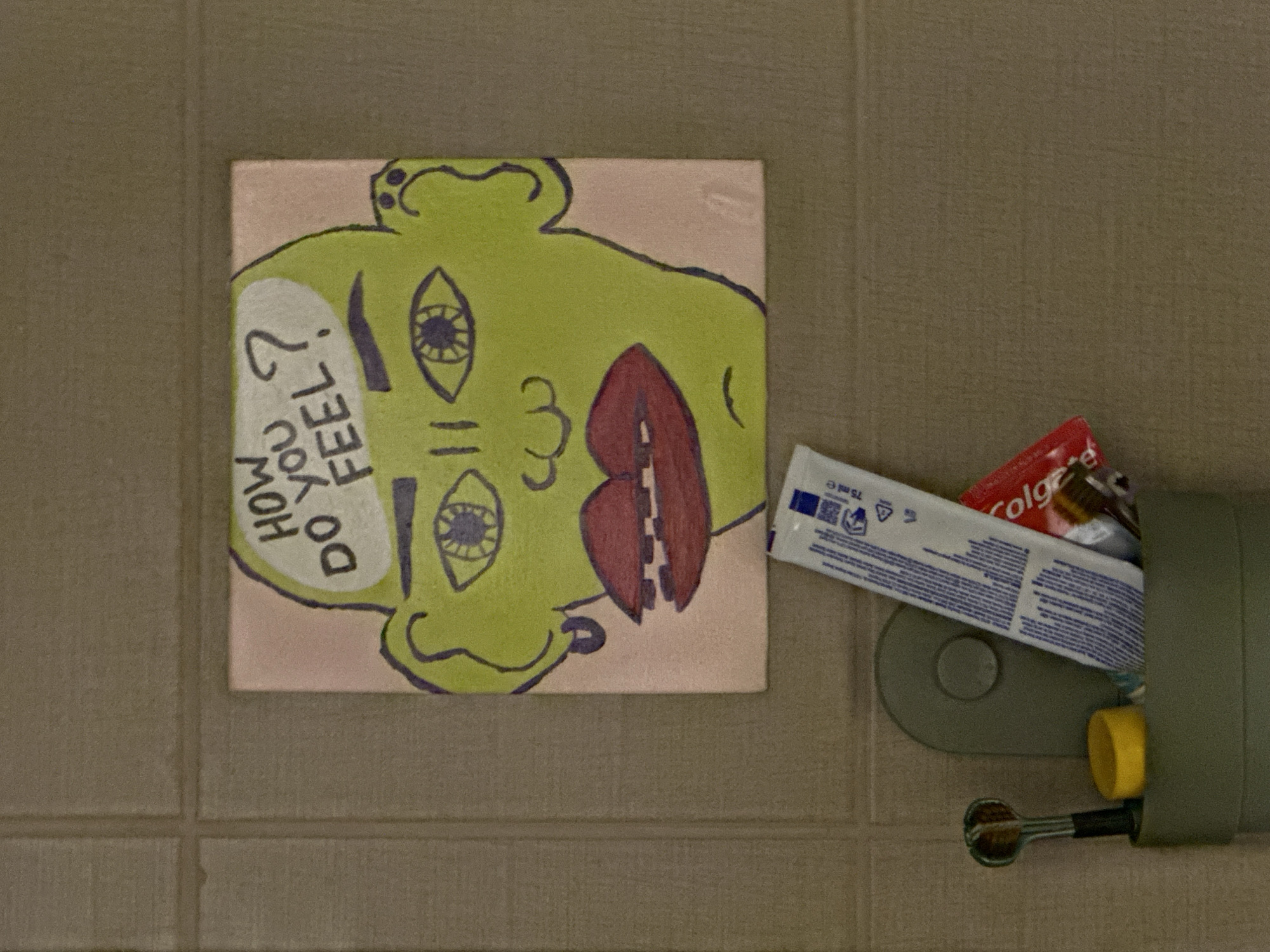 A square canvas painting of a stylized green head with large, expressive eyes and full red lips is affixed to a tiled wall. The head is bald, and text within a thought bubble above it reads "HOW DO YOU FEEL?". The painting is positioned above a gray plastic organizer containing a tube of toothpaste, a red package that appears to be toothpaste as well, and a toothbrush. The background is a grid of textured, taupe-colored tiles. This scene is located in Budva, Montenegro.
