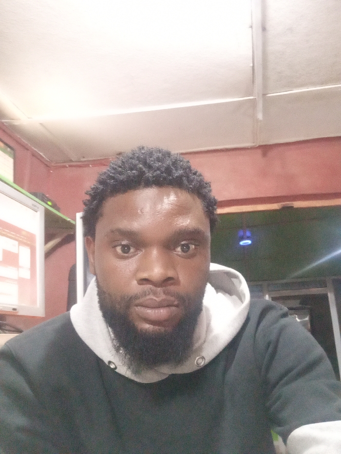 A close-up shot showcases a dark-skinned man with short, curly hair and a full beard. He is wearing a dark hoodie with a light gray hood lining. His expression is neutral, with direct eye contact. The background is a dimly lit room with a computer monitor visible on the left side. The lighting in the image creates a slightly grainy and muted color palette.