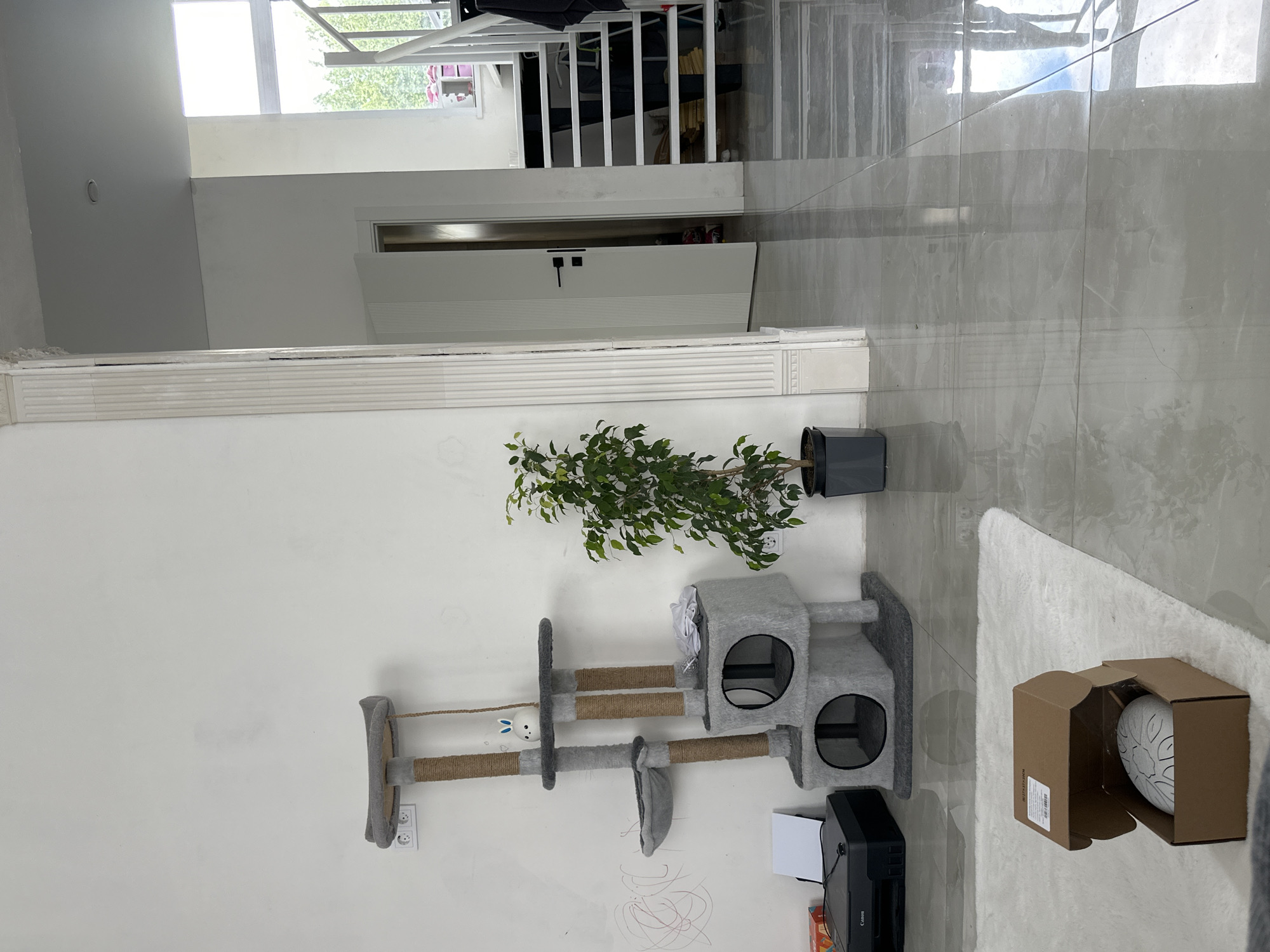 The image depicts an indoor scene within a residential or commercial building in Unknown, Unknown. The primary visual elements are bright white walls and a highly reflective light grey tiled floor.
On the left, a white wall displays faint red, scribble-like markings. A black Canon printer sits on the floor beneath a white double electrical outlet. A multi-level cat scratching post, composed of grey fabric platforms and sisal rope-wrapped pillars, stands against this wall. It features two enclosed compartments at its base, an oval-shaped hanging bed, and multiple elevated platforms. A small white spherical figurine with blue-tipped bunny ears is placed on an upper platform.
Towards the center, a leafy green plant in a dark grey pot is positioned next to a white decorative pilaster column. The column, exhibiting vertical fluting, appears to be partially integrated into the wall. Beyond this column, a white door with vertical grooves and a black handle is ajar, revealing a darker room beyond. To the right of the door, a white open-riser staircase with a white metal handrail ascends. Dark fabric items are draped over the staircase railing. A window at the top of the stairs shows green foliage outdoors.
In the foreground, a white shaggy rug lies on the tiled floor. An open brown cardboard box, containing a white tongue drum with black geometric designs, rests on the rug. The reflective tiled floor surface clearly displays reflections of the ceiling, the door, and the lower portion of the staircase.