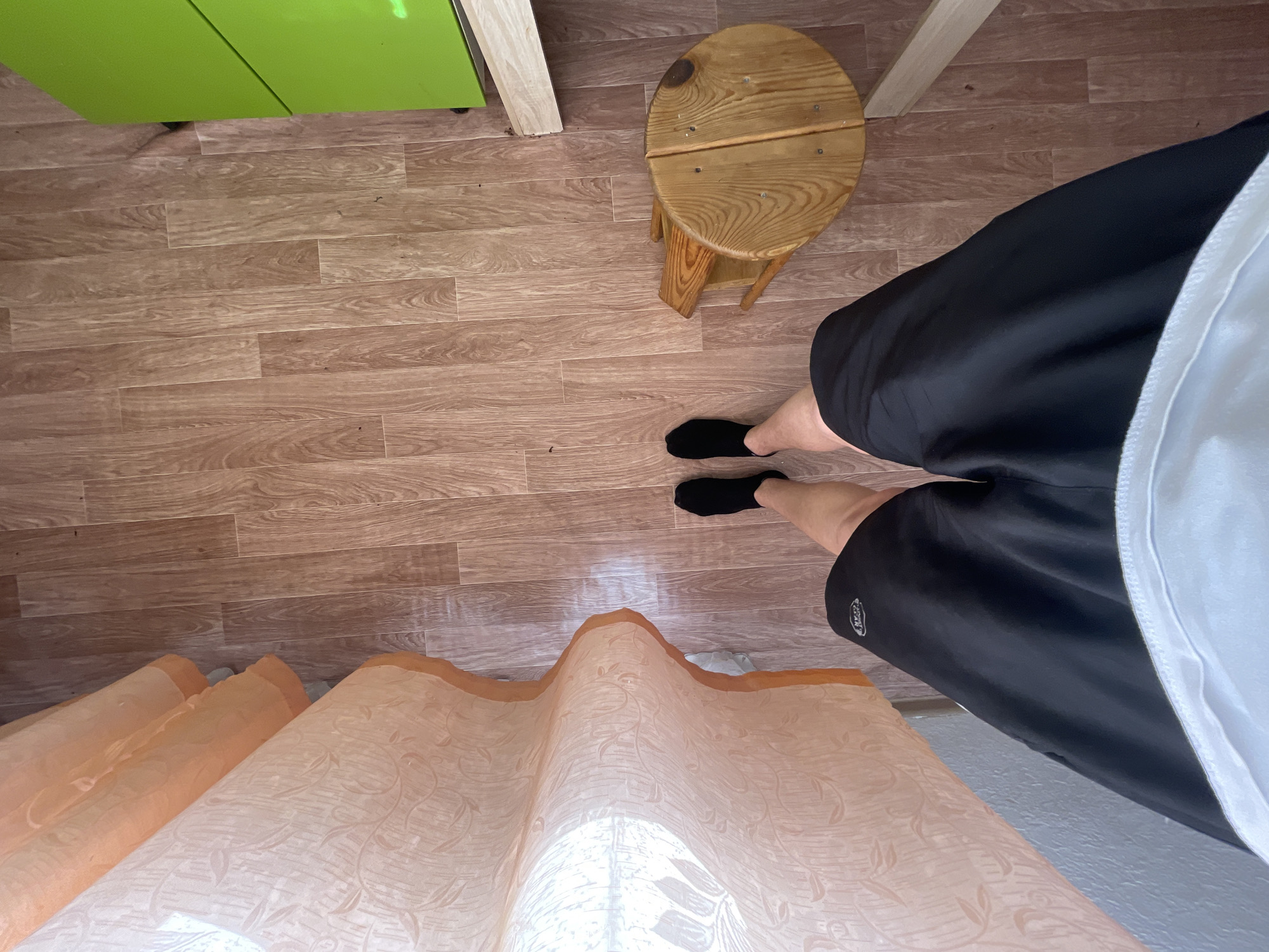 The image shows a first-person perspective of a person standing on a wood-patterned floor. The person is wearing black shorts and black socks. To the left, there is a sheer orange and white curtain with a floral pattern. To the right, a small, circular wooden stool is visible, and further to the right, a bright green surface is partially visible, likely part of a cabinet or appliance. The scene appears to be indoors, possibly in a domestic setting. The location is identified as Красные Баки, Russia.