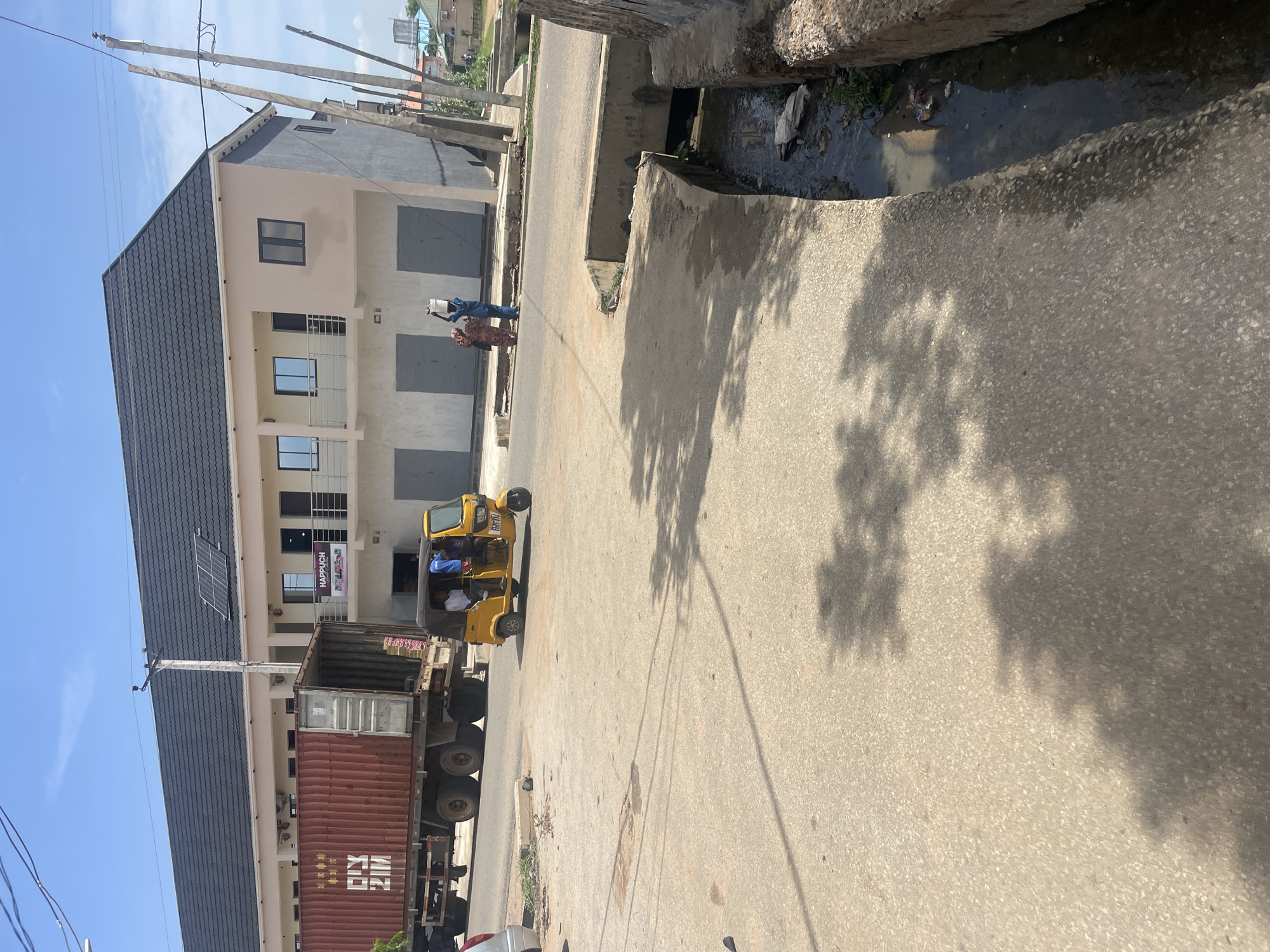 This media file captures a street scene in Jimeta, Nigeria, likely during the daytime under a clear sky. The image is oriented vertically, showing a street lined with buildings and a drainage channel.
In the foreground, a large truck carrying a shipping container is parked on the right side of the road. Next to it, a yellow auto-rickshaw, also known as a "Keke Napep," is stopped, with several passengers visible inside. The rickshaw has text on its side, but it is not clearly legible.
Further down the street, two individuals are walking. One is carrying a white bucket on their head, and the other is dressed in a patterned fabric. Behind them, a multi-story building with a dark-tiled roof and light-colored walls stands prominently. This building features several windows and balconies, and what appears to be a sign with the text "HAPPULCH" and an image of a woman. A solar panel is affixed to the roof of this building.
The street is paved with asphalt, and a concrete drainage channel runs alongside the road, containing murky water and some debris. The shadows of trees are cast across the road and the drainage channel, suggesting the presence of nearby foliage and the time of day. The overall scene depicts a typical urban street environment in Nigeria, with everyday activities of transportation and pedestrian movement.