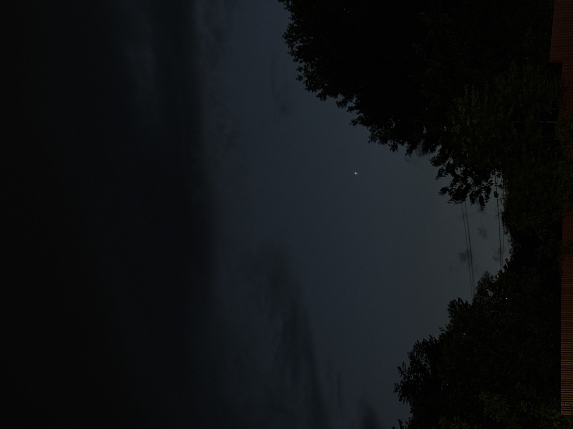 The image shows a dark night sky above a silhouette of trees and a wooden fence. A single bright star is visible in the upper right quadrant of the sky. The clouds are dark and heavy, suggesting a potentially overcast or stormy night. The overall impression is one of a quiet, rural or suburban landscape at night. There are no people or animals visible in the image. The scene is static and serene. It is impossible to determine the specific location within Mikhaylovsk, Russia from the provided image.
