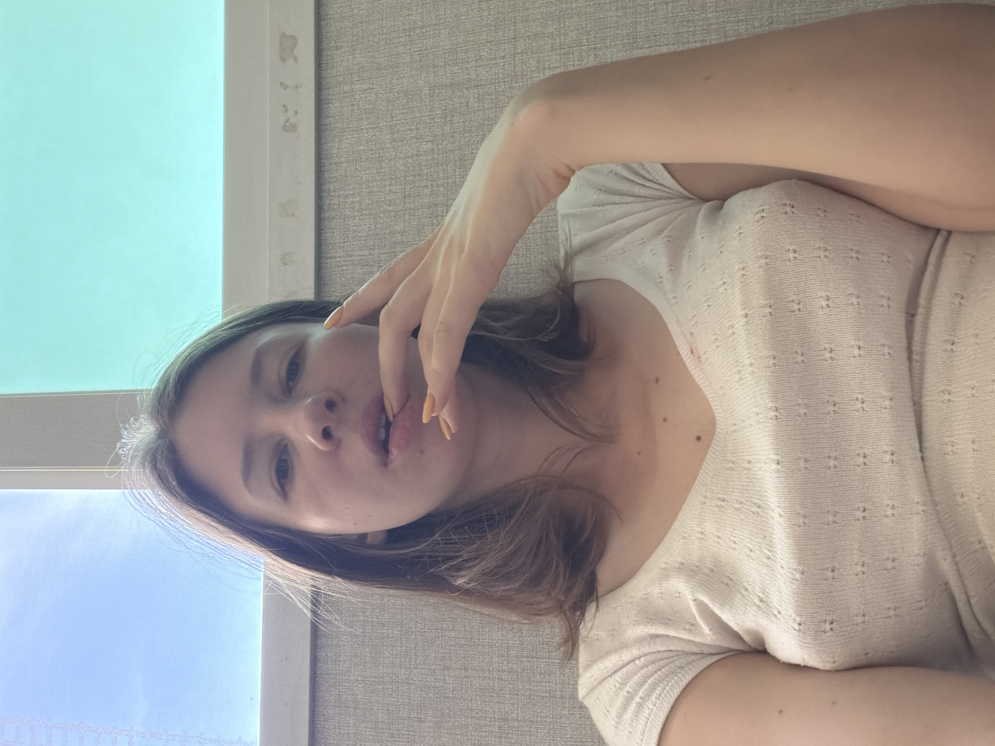 A young Caucasian female is pictured from the chest up, looking directly at the camera. She has medium-length brown hair, and her left hand is raised to her mouth. She is wearing a white V-neck t-shirt with a textured pattern. The background consists of a wall with a textured grey wallpaper and a window with a blue sky visible through the top pane and a teal-colored pane below. The image appears to have been taken indoors, likely in a residential or office setting, with natural light coming from the window. There are no visible landmarks or specific details that would indicate a precise location within Dongducheon-si, South Korea, beyond the general assumption of the setting.