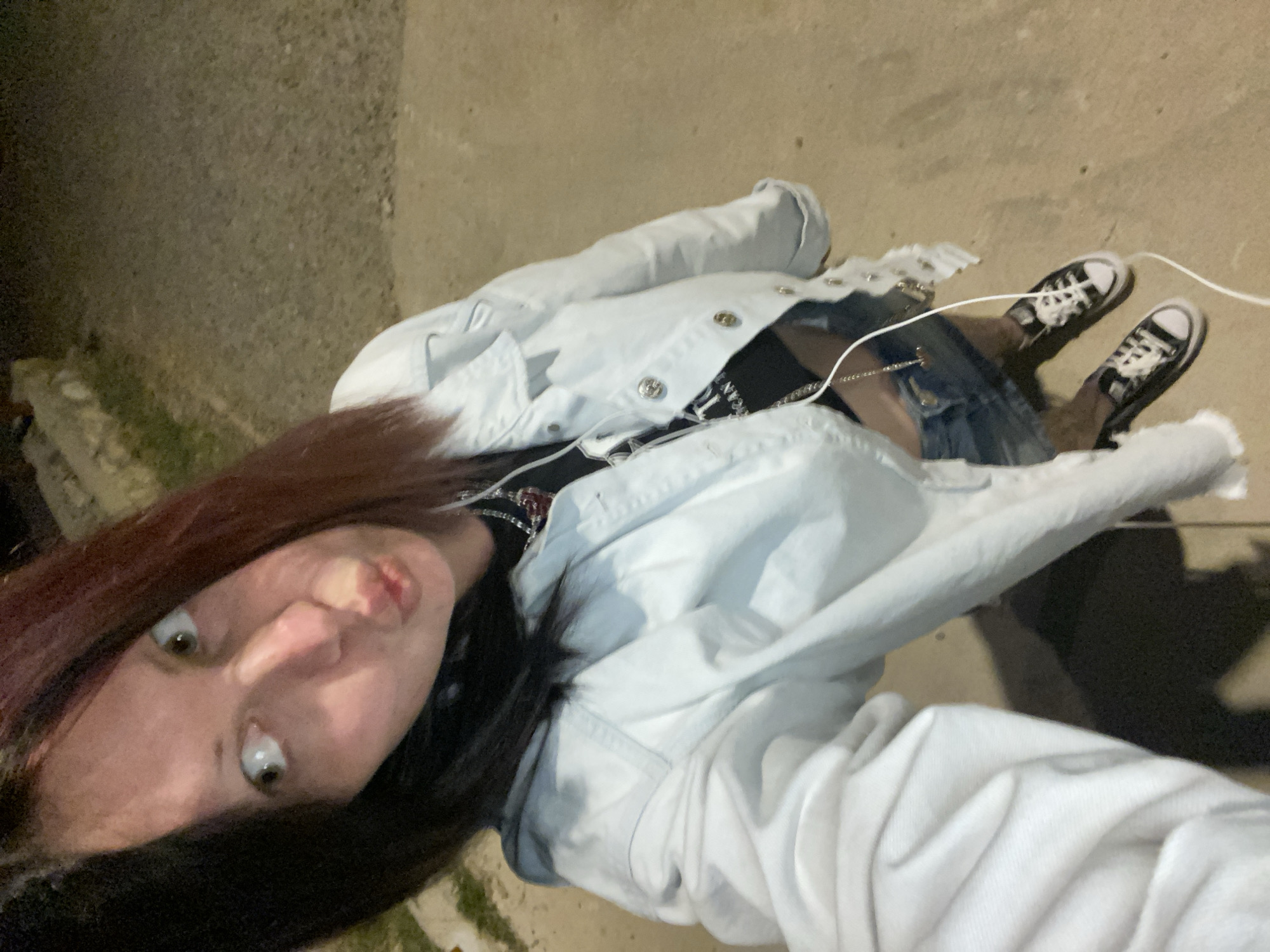A selfie captured at dusk. The subject, a young person with auburn and black hair, is facing the camera with pursed lips. They are wearing a light-colored denim jacket over a dark top with visible lettering, a denim skirt, and black-and-white Converse sneakers. A white earphone wire is visible. The background is a concrete or asphalt surface. The location is not identifiable as Kunje, Montenegro, from visual information.