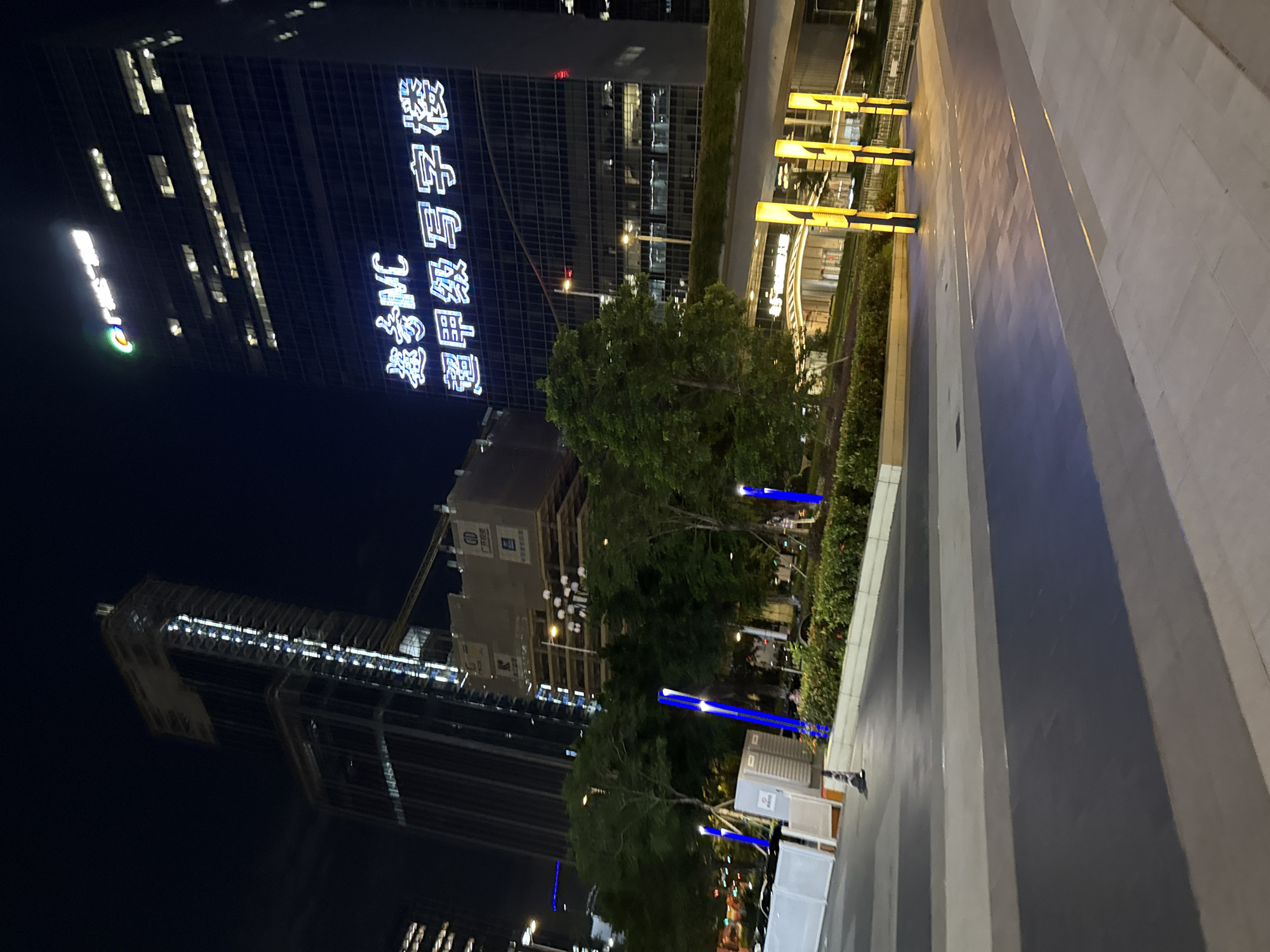 This image depicts a nighttime view of a modern urban landscape in Huangpu District, China. In the background, two prominent skyscrapers dominate the skyline. The taller building on the left appears to be under construction, with visible scaffolding and unfinished sections. The building on the right is a completed high-rise with illuminated windows and a large, bright sign displaying Chinese characters and "MC" in English. The sign reads "嘉信MC 超甲级写字楼," which translates to "Jiaxin MC Super Grade Office Building."
In the foreground, a paved walkway with distinct patterns of dark gray, light gray, and blue sections leads the eye towards the buildings. To the left of the walkway, there are white barricades and portable restroom units. Several tall, slender, blue-lit poles are strategically placed along the edge of the walkway, providing ambient lighting. Lush green trees and manicured bushes are visible between the walkway and the buildings, adding a touch of nature to the urban environment. Further into the mid-ground, a series of tall, yellow-lit cylindrical structures stand out, illuminating the area beneath an elevated walkway or bridge. The overall scene conveys a sense of modernity and urban development.