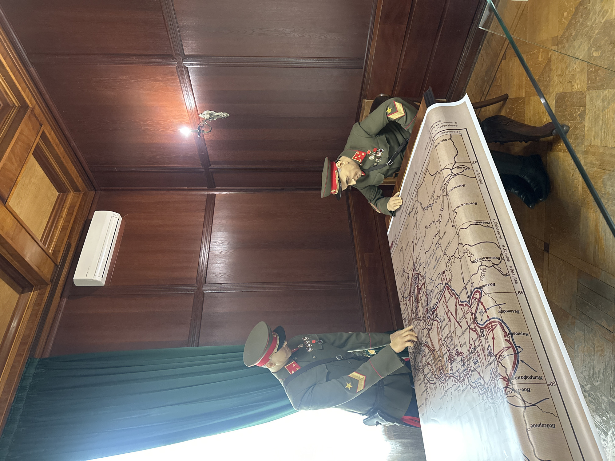 This is a photograph taken inside a room in the city of Sochi, Russia. Two mannequins dressed in Soviet military uniforms are positioned around a large, detailed map. The mannequins appear to be studying the map, suggesting a historical or educational context. The room has a dark wood-paneled wall, an air conditioning unit, green curtains, and a dark wooden floor, indicating the setting may be a museum or historical exhibit. There is a glass barrier at the right side. The map contains handwritten labels in Russian. It contains the name of different locations (e.g., “Belovodsk”, “Voroshilovgrad” and “Pavlovskaya”).
