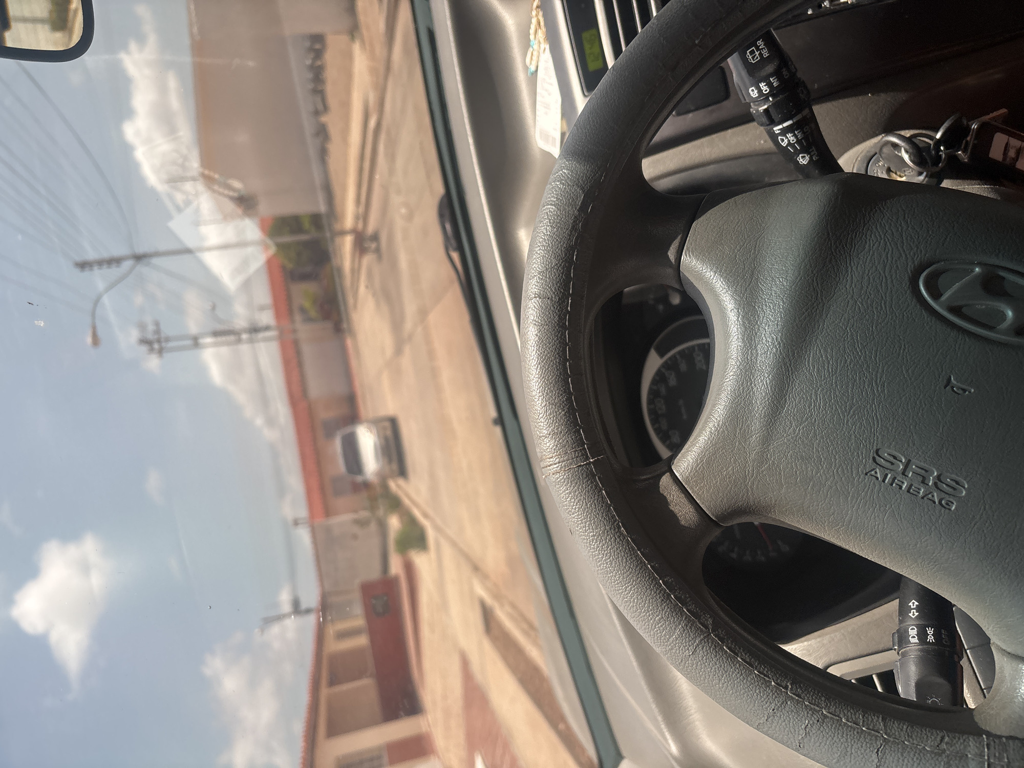 The image, taken from the interior of a car, shows a view through the windshield. In the foreground, the gray leather-wrapped steering wheel is prominent. To the left of the steering wheel, a turn signal stalk with symbols for headlights and turn signals is visible. To the right of the steering wheel, a wiper control stalk with settings for "OFF", "INT", "LO", and "HI" for the front wipers, and "REAR" for the rear wiper is present. A car key is inserted into the ignition.
The scene outside the car appears to be a street in a residential area. Houses with orange tiled roofs line the street, and a light-colored car is parked further down. Power lines and streetlights are visible overhead. The sky is blue with scattered white clouds, suggesting it is daytime and likely sunny. The overall environment suggests a typical urban or suburban street scene.