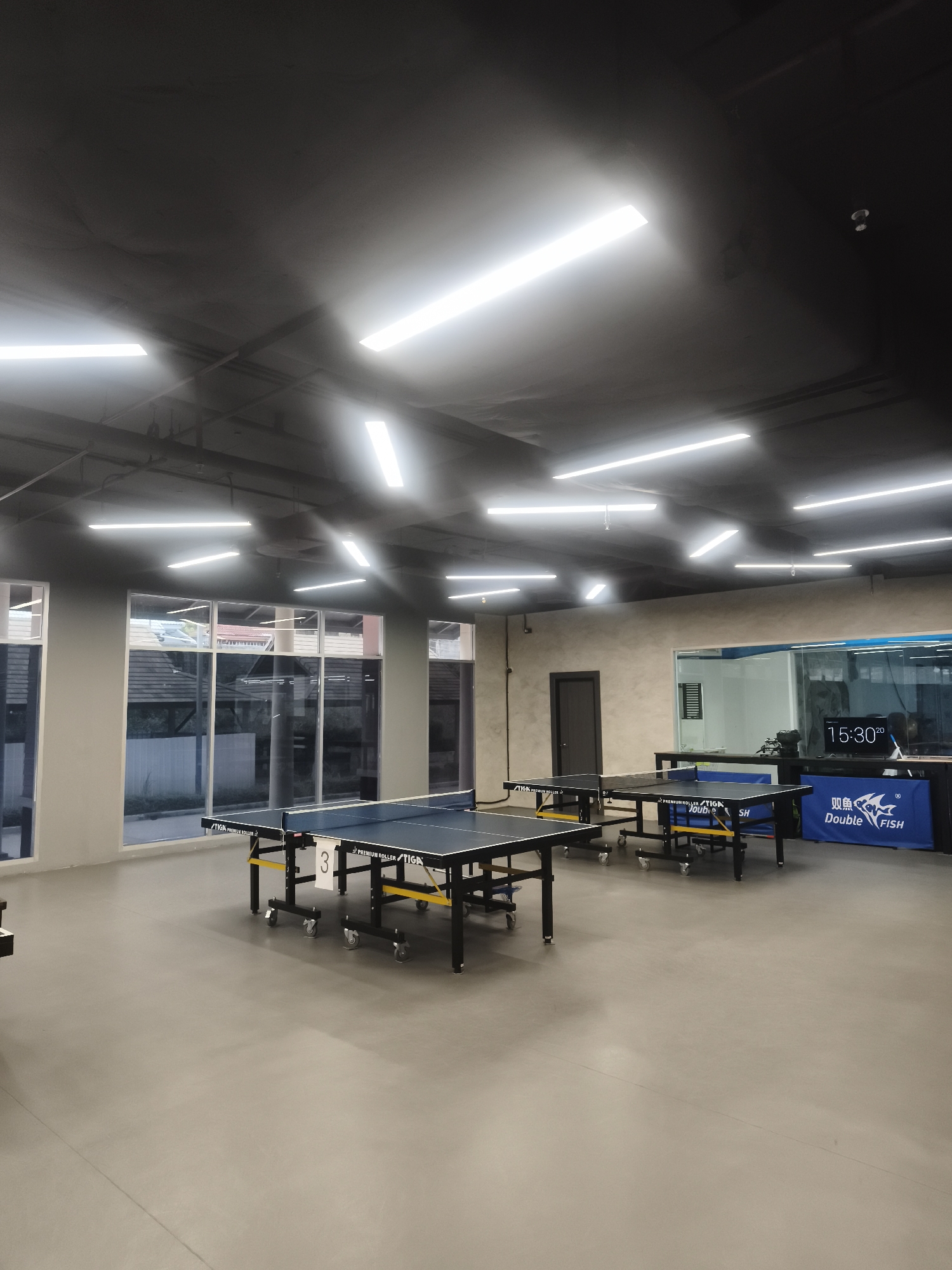 The image depicts a well-lit indoor space, possibly a recreation or sports hall. The space is dominated by three ping pong tables with the brand "STIGA Premium Roller" visible. One of the tables is marked with a "3". A banner with the logo "Double Fish" is behind one of the ping pong tables, along with a digital clock that shows the time 15:30. The hall is illuminated by several bright, linear light fixtures arranged across the ceiling. The walls are neutral in color, and large windows can be seen on one side of the hall.
The possible location of the scene is บ้านบางรักษ์, Thailand.
