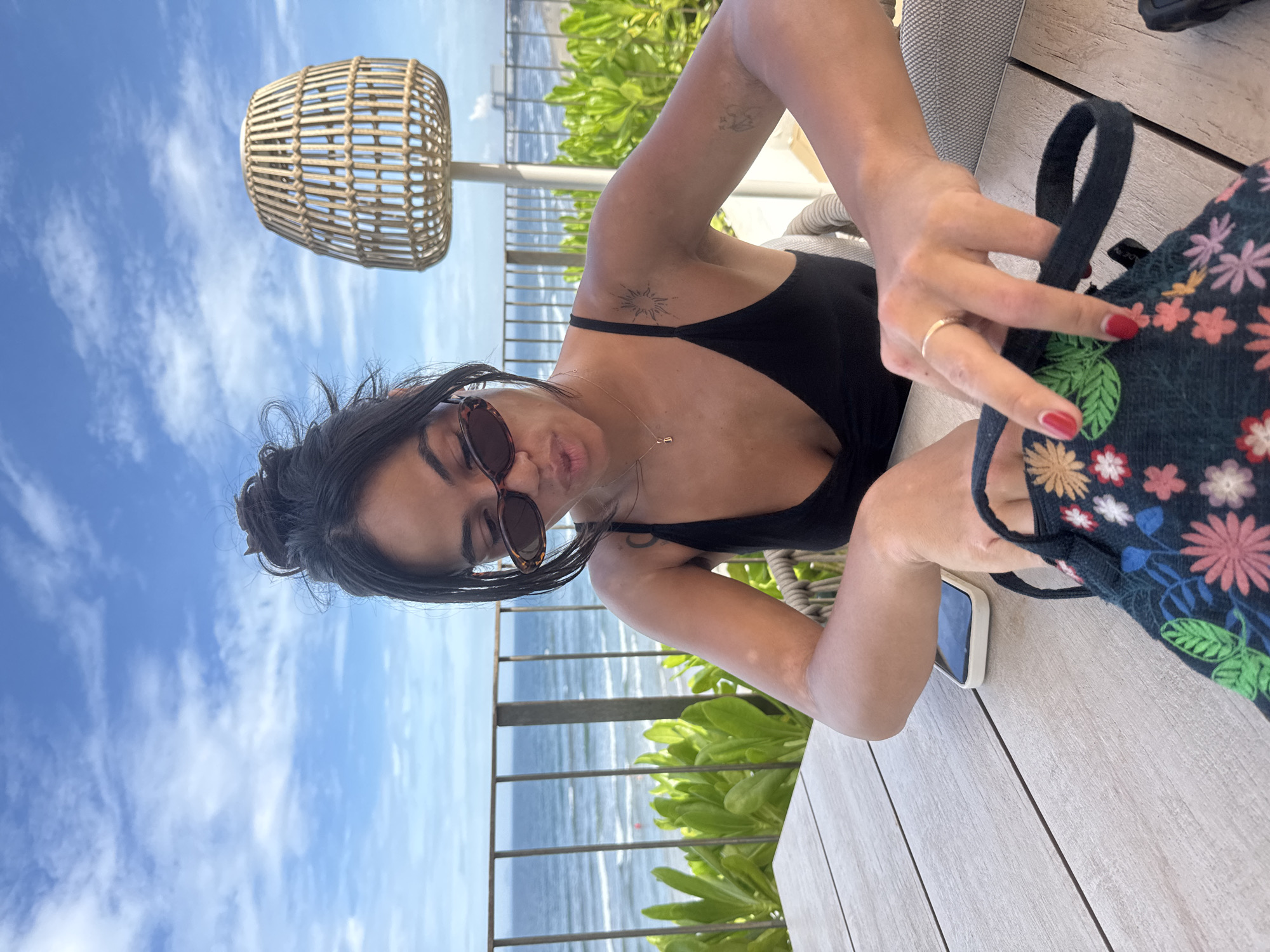 A woman sits at a light-colored wooden table with a floral bag, her phone, and a woven lamp visible. She wears a black top and sunglasses. The backdrop shows a metal railing, green foliage, the ocean, and a blue sky. This photograph was likely taken at a beachfront establishment in Đà Nẵng, Vietnam.