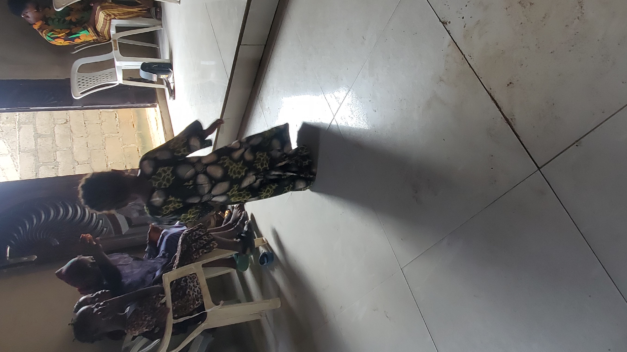 This image captures an indoor scene in Jalingo, Nigeria, featuring multiple individuals and simple furnishings under natural lighting.
In the foreground, a young child, viewed from behind, walks rightward across a light-colored tiled floor, casting a pronounced shadow ahead. The child wears a long-sleeved dress with a dark background patterned with white and green elements. The tiled floor exhibits visible grout lines and some minor discoloration/dirt.
To the left, three individuals are seated on white plastic chairs. The leftmost individual, possibly a child or young adult, wears a leopard-print top and faces right. Next to them, a person in dark attire is partially obscured. The third person in this group, an adult, wears a purple head covering and top, and appears to be holding a small dark object (possibly a remote) towards a dark, fan-like unit mounted higher on the wall. Bare feet and several pairs of sandals/slippers (including blue and black ones) are visible on the floor beneath their chairs.
In the background, a brightly lit doorway reveals an exterior wall constructed of light-colored concrete blocks. To the right of the doorway, another individual sits on a white plastic chair, facing left and looking downwards. This person wears a patterned top with yellow, orange, and green elements, and striped trousers. A small black bag or case rests on the floor near their chair.
The scene is lit by strong natural light entering through the doorway, creating bright reflections on the polished floor tiles and casting shadows. The interior walls appear plain and light-colored.