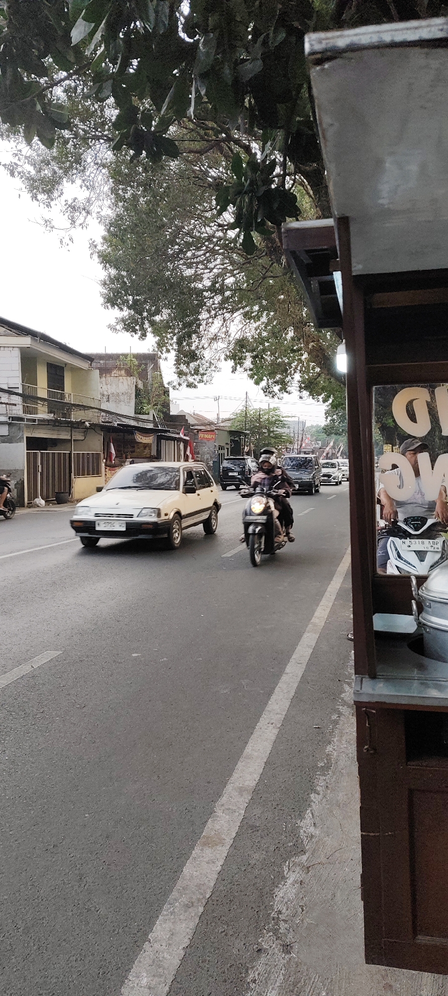 A vertical, medium shot captures a street scene in Malang, Indonesia, likely during daylight hours. The foreground features the edge of a food cart with a dark brown wooden exterior. The cart has a light gray metal lid on top and appears to be selling food, possibly steamed items, given the visible stacked containers. A man in a white shirt and a gray cap is partially visible, seated on a white scooter with a license plate reading "N 5318 ABP".
The street itself is asphalt with a faded white lane marker. Several vehicles are present: a white hatchback car with a license plate reading "W 2724 HD" is moving in the left lane. A scooter with a rider wearing a black helmet and jacket is in the right lane, moving in the same direction as the car. Further down the street, other vehicles, including dark-colored cars and possibly a truck, are visible.
On the left side of the street, there are buildings with two stories. The lower levels appear to be commercial spaces, with doors and windows visible. Some of these buildings have awnings or overhangs. Indonesian flags are displayed on some of the buildings. Trees with dense green foliage overhang the street, casting shadows and contributing to the natural environment of the scene. The sky is overcast, suggesting a cloudy or hazy day. The overall impression is of a typical urban street in Indonesia with moderate traffic.