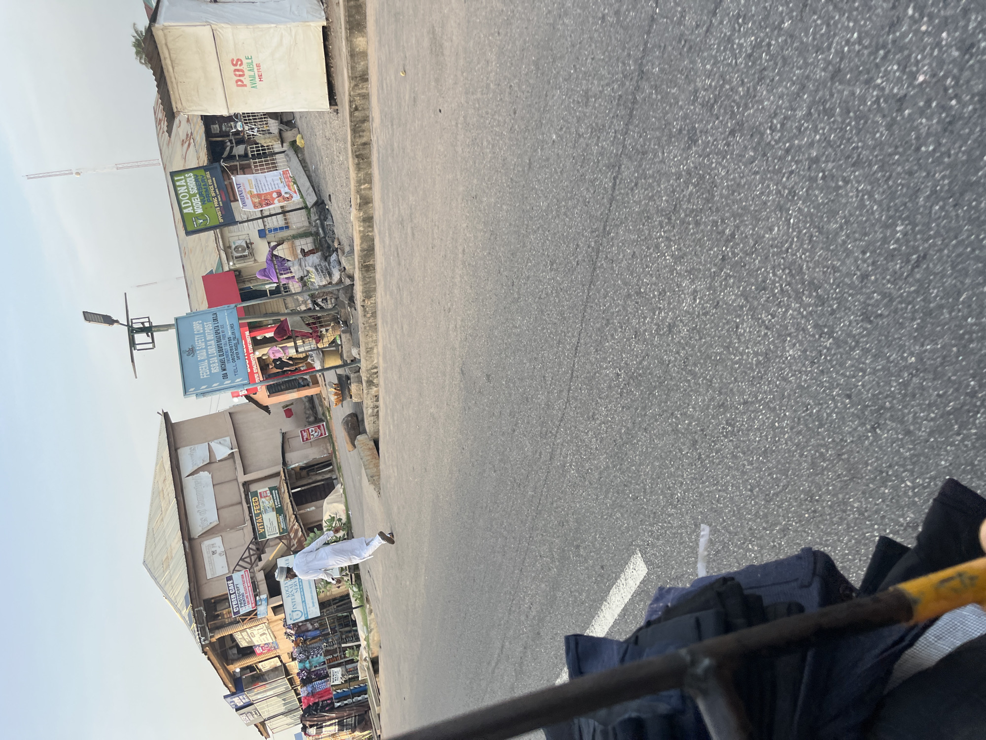 An outdoor, eye-level, medium shot shows a man in a white robe and cap walking away from the camera on a paved street. The man is in the middle of the frame. To his left are various shops with signs and displays of clothing. One sign reads "VITAL FEED". To the right of the man are more shops and a large white tent with "POS AVAILABLE HERE" written on it. The street is asphalt with a white line dividing the lanes. The background shows a building with a tin roof and more signs. The sky is clear and pale blue.