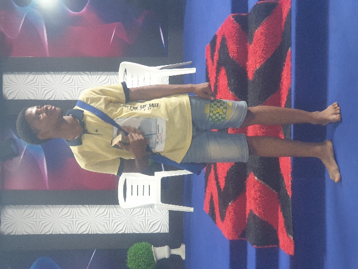 One young Black male stands barefoot in an indoor setting in Onitsha, Nigeria. He wears a short-sleeved yellow polo shirt with dark blue trim, light blue denim shorts with a green, white, and yellow checkered pattern on the right thigh, and a blue strap across his left shoulder. He holds a dark-colored book with his left hand against his chest, and a white identification tag with text and an image is visible on his shirt. The individual is oriented slightly to his right, looking upwards and away from the camera. He stands on a blue carpeted floor in front of a two-tiered blue carpeted stage or platform, which is adorned with a shaggy red and black chevron-patterned rug on its steps. In the background, two white plastic chairs are visible on the stage level. To the far left, a white planter holds a green artificial plant. The backdrop wall features a textured white panel with a geometric pattern on the left and a dark panel with flowing, multi-colored (blue, pink, white) abstract lines on the right.