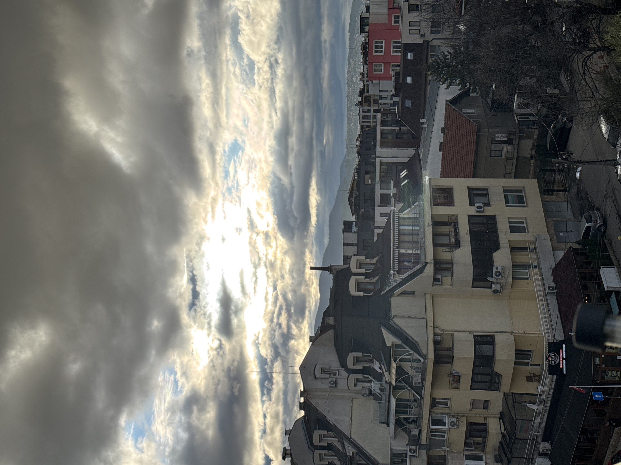 The image shows a view of the city of Sofia, Bulgaria, under a cloudy sky with the sun breaking through. A variety of buildings are visible, including residential buildings, some with red roofs. A red building can also be seen. Most buildings in the foreground appear to be residential apartments. A small sign is visible at the corner of a building. The street is visible and shows cars. Mountains can be seen in the distance.