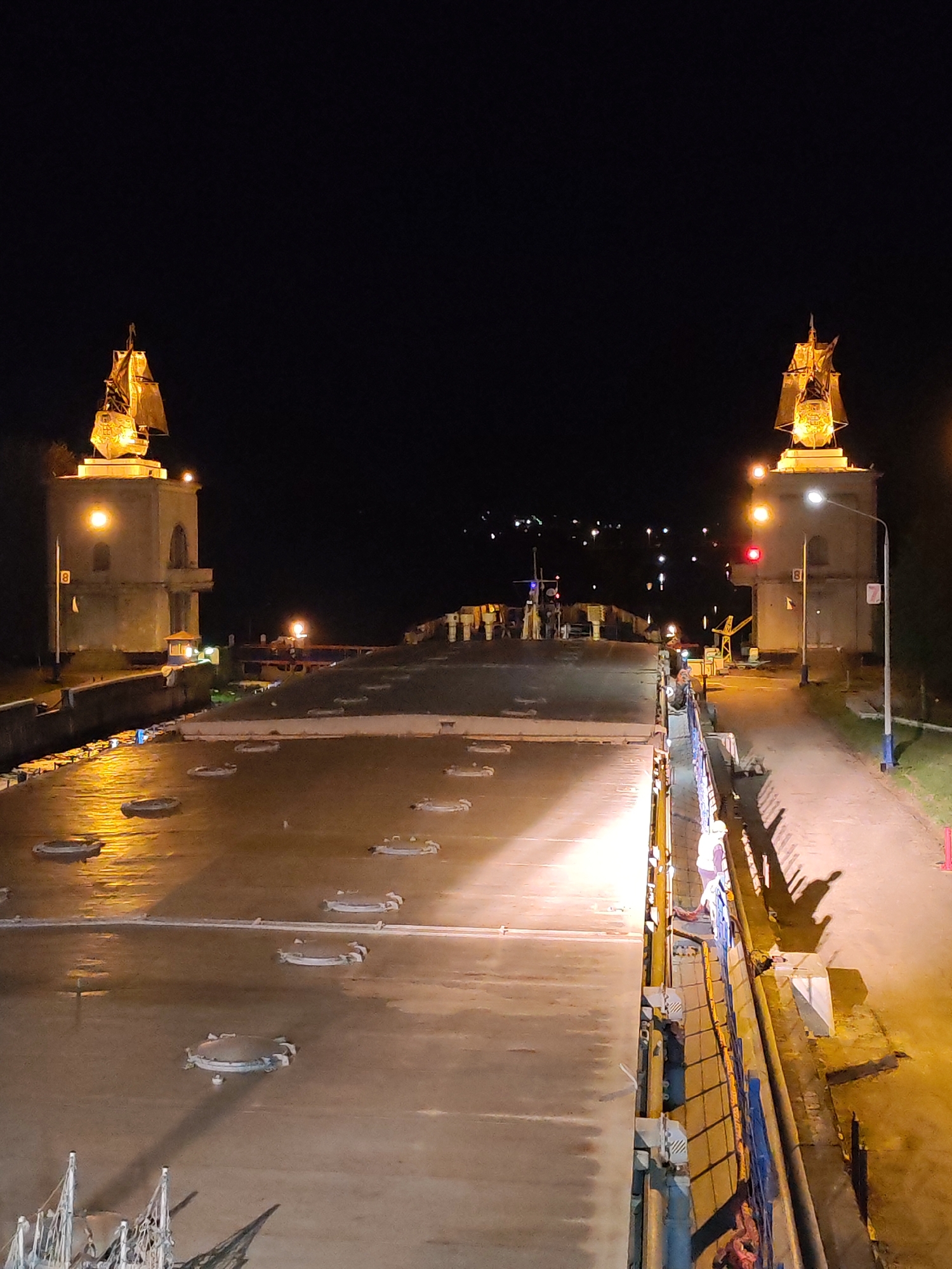 Captured at night, the scene depicts a water-filled lock on the Moscow Canal in Yakhroma, Russia. Flanking either side of the lock are identical structures, each topped with illuminated model ships. A pathway runs alongside the lock on the right. A person wearing a light-colored jacket is visible standing next to the railing along the pathway. The water in the lock is dotted with numerous circular objects, reflecting the artificial light sources. The background features the dim glow of city lights in the distance.