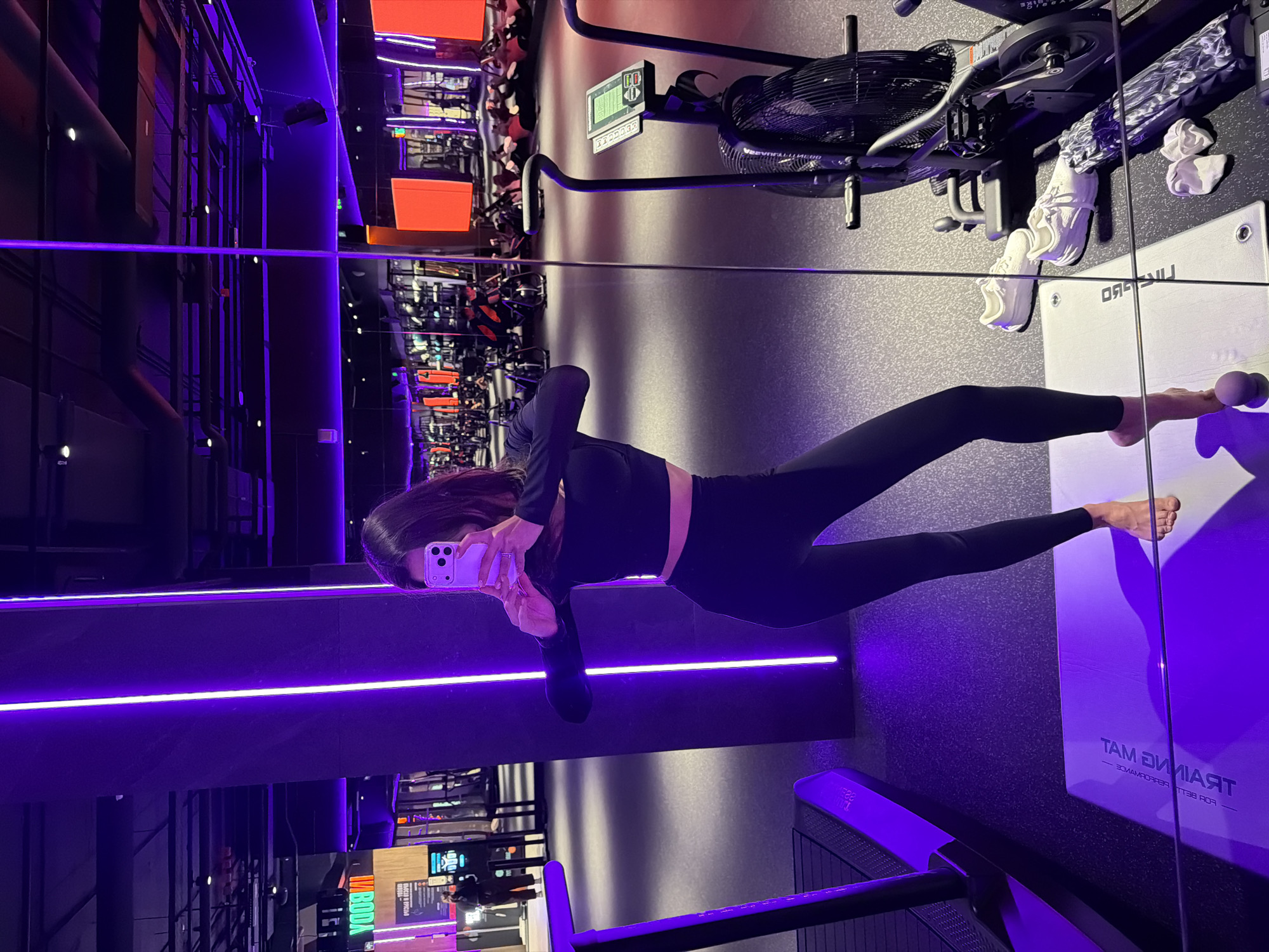 A mirror selfie captures a woman posing in a modern fitness studio in Krasnoyarsk, Russia. The image is oriented vertically, showing the woman's full reflection in a large mirror or reflective surface. She is dressed in black athletic wear, including a long-sleeved crop top and leggings, with her bare feet visible. Her face is obscured by the smartphone she holds up to take the picture.
The gym features a contemporary, dark aesthetic, predominantly lit by prominent purple LED strip lights that run horizontally along a wall structure, creating a vibrant, yet dim, atmosphere. Various fitness equipment is visible in the background and foreground, including an air bike with a large fan, white athletic shoes, and a foam roller. A white workout mat is positioned on the floor, displaying the text "TRAINING MAT" and "FOR BETTER PERFORMANCE" in reverse due to the reflection. Other gym apparatus and display screens, with partially visible text like "INBODY," are blurred in the background reflections. The artificial lighting suggests the photograph was taken during an evening or nighttime workout session.